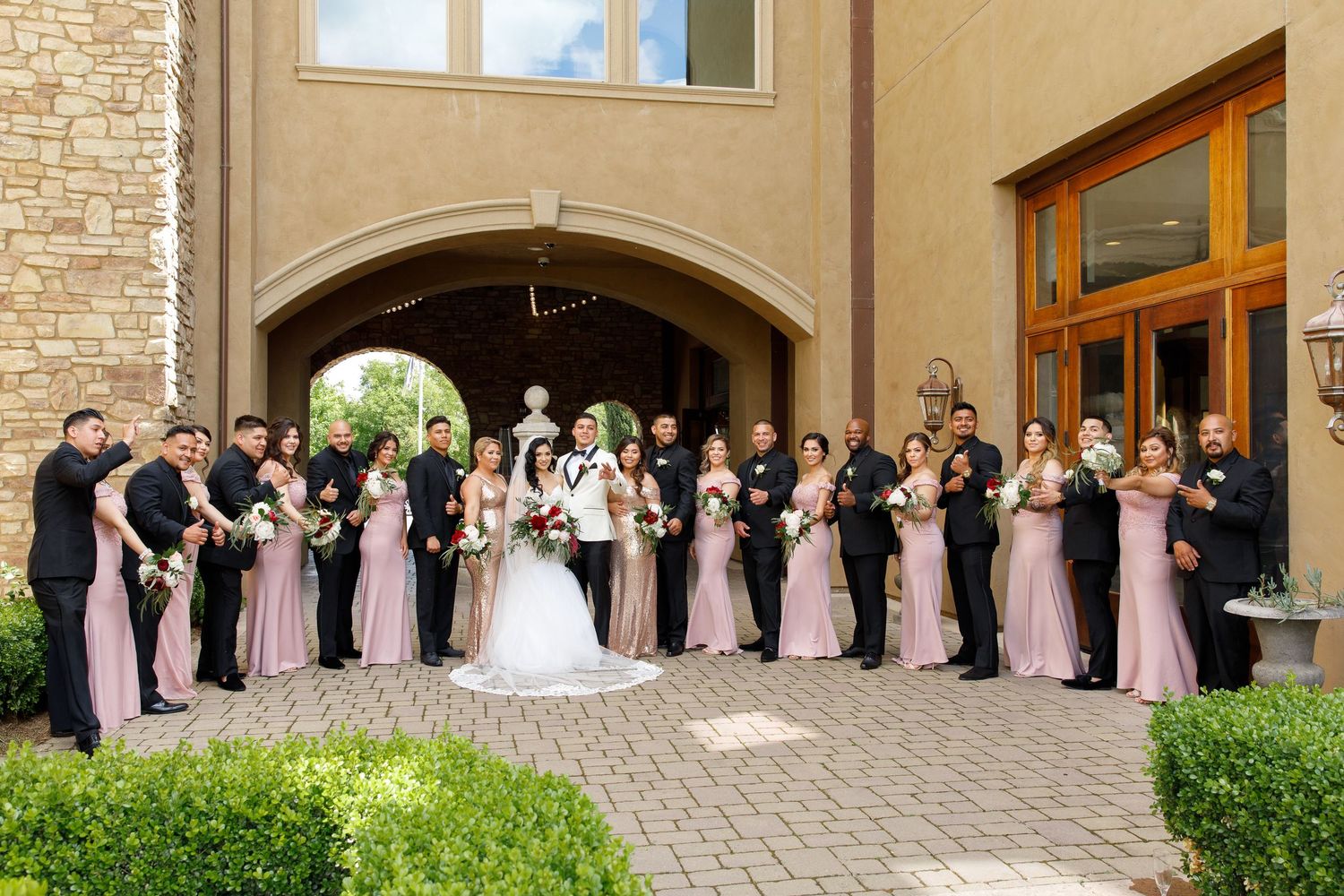 Large wedding party in pink dresses poses under stone archway at elegant venue during sunset.
