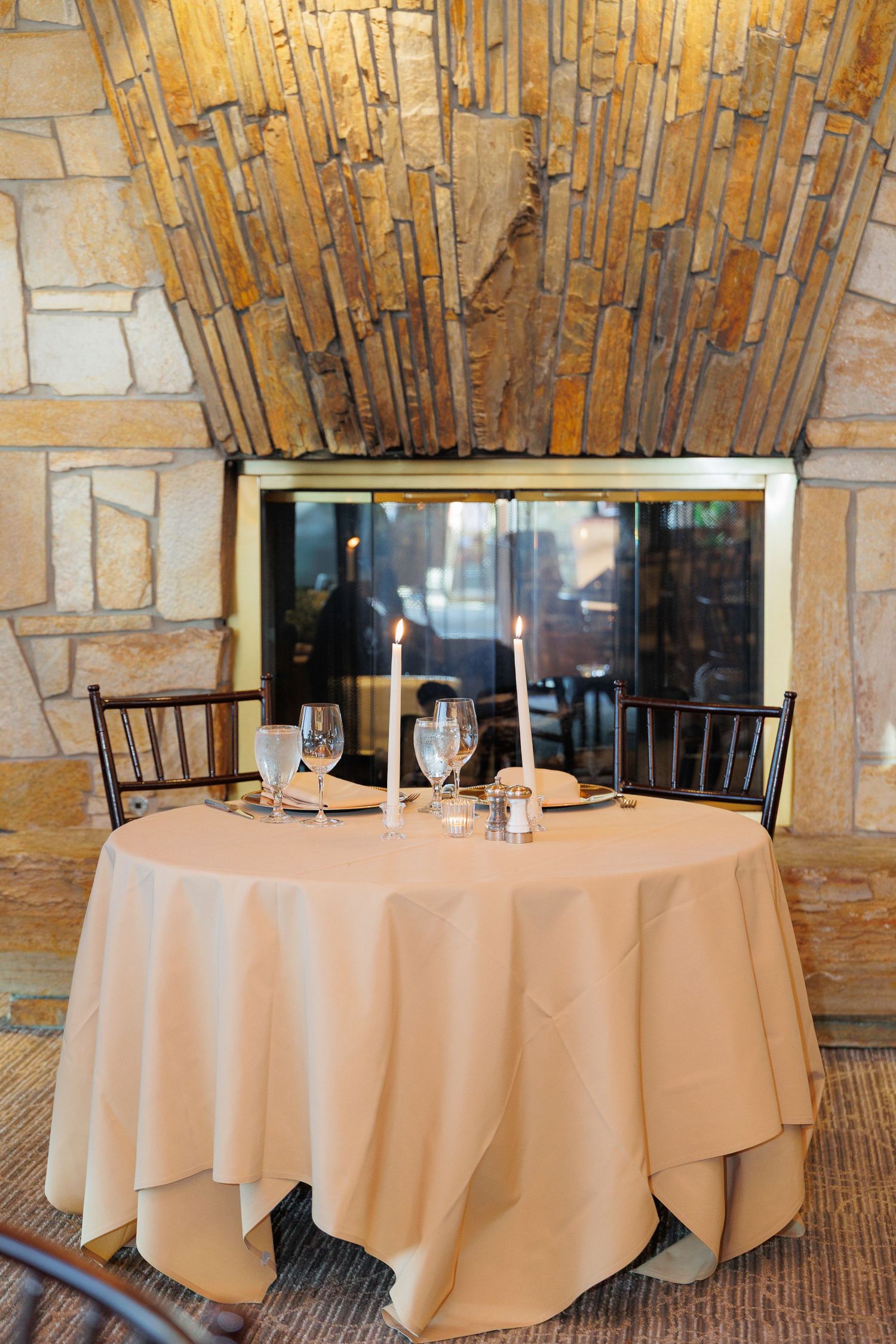 Elegant restaurant table setting with beige tablecloth and wine glasses against stone wall with bamboo decor.