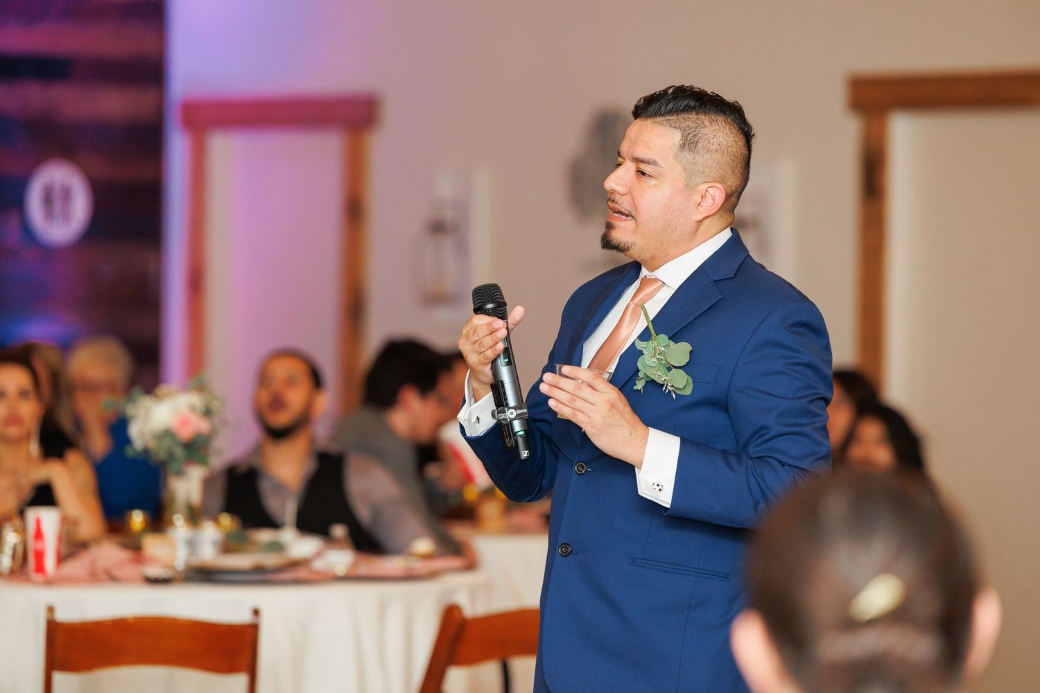 A person in a blue suit gives a speech at a wedding reception while holding a microphone.