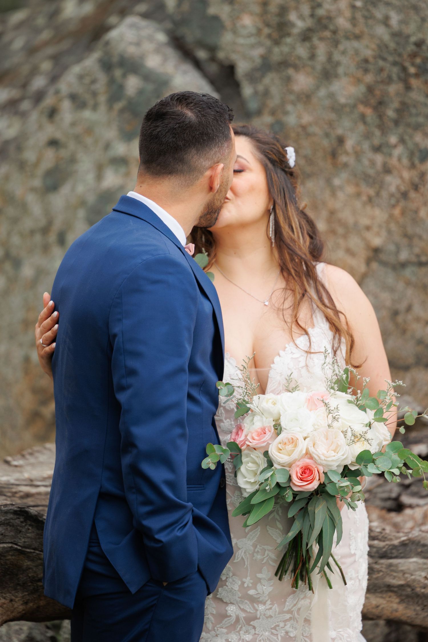 A bride in a white lace gown and groom in navy suit embrace while holding a blush and cream floral bouquet.