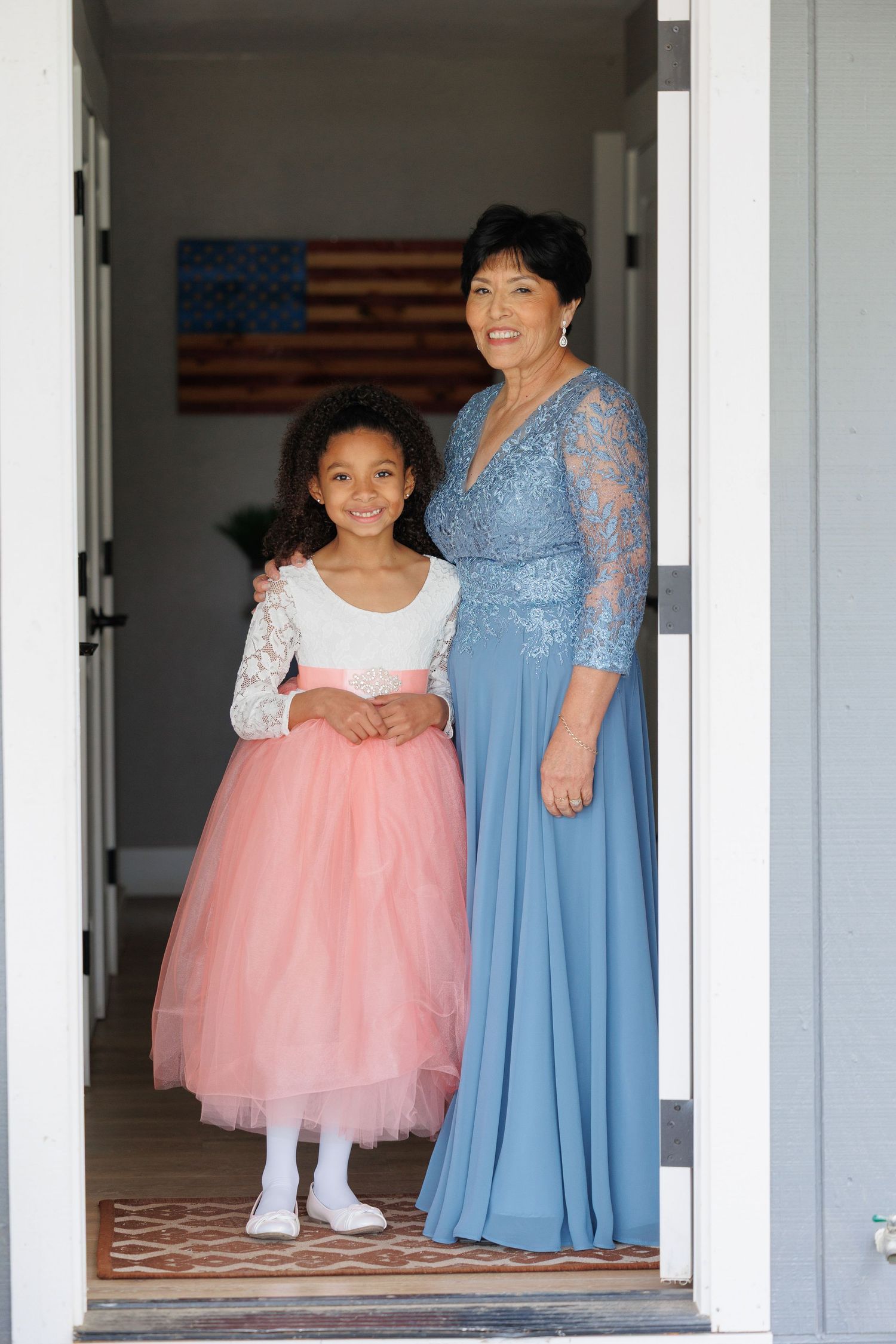 A young girl in a pink tulle dress stands with someone wearing a blue gown in a white doorway.