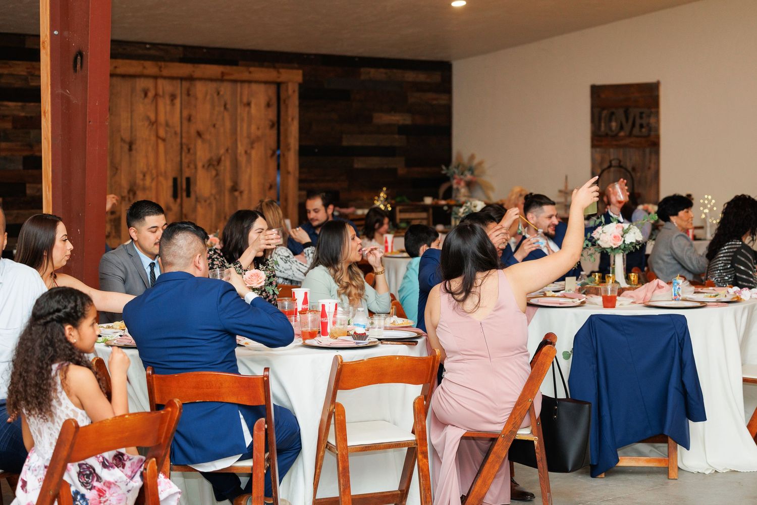 Wedding guests enjoy dinner at round tables with white tablecloths in a rustic venue with wooden walls.