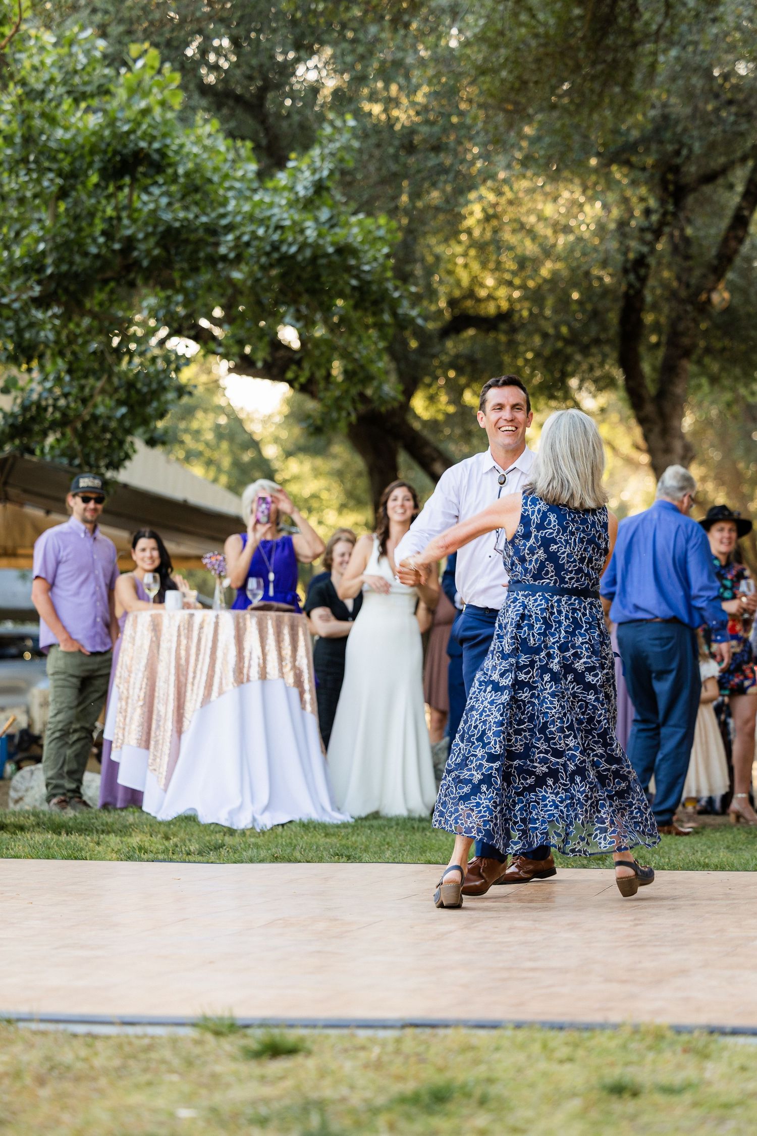 Guests gather at an outdoor wedding reception under shady trees with white cocktail tables and blue attire.