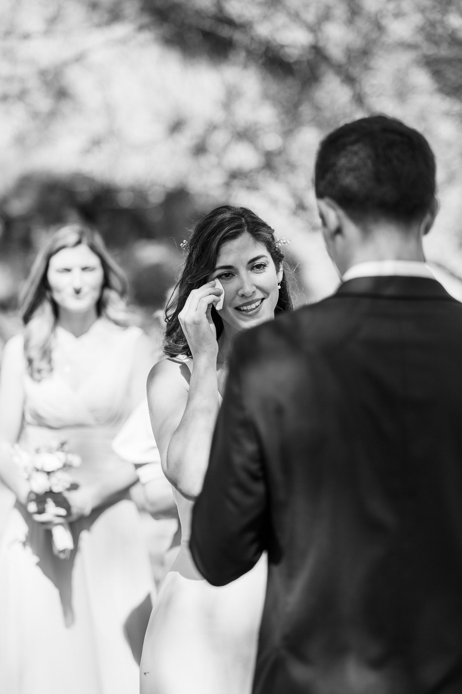 Black and white wedding photo showing emotional moment during outdoor ceremony with bride wiping away tears.