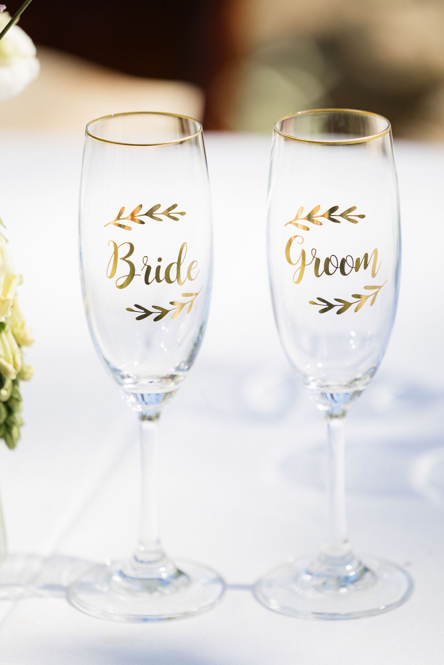 Two elegant champagne flutes with gold lettering reading 'Bride' and 'Groom' decorated with delicate leaf designs.