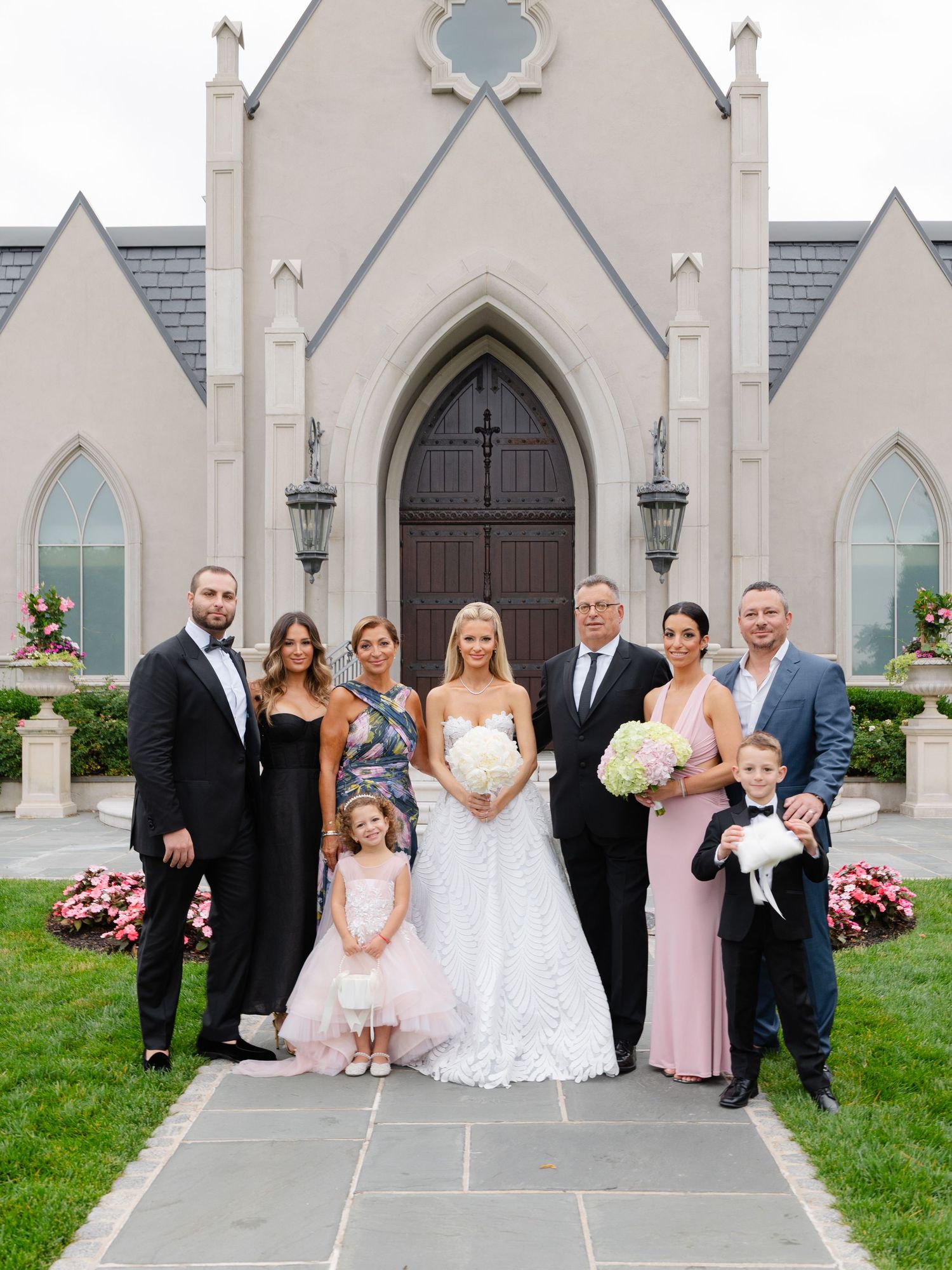 Wedding party poses in front of a Gothic church with arched doorways and pink hydrangeas blooming in the garden.