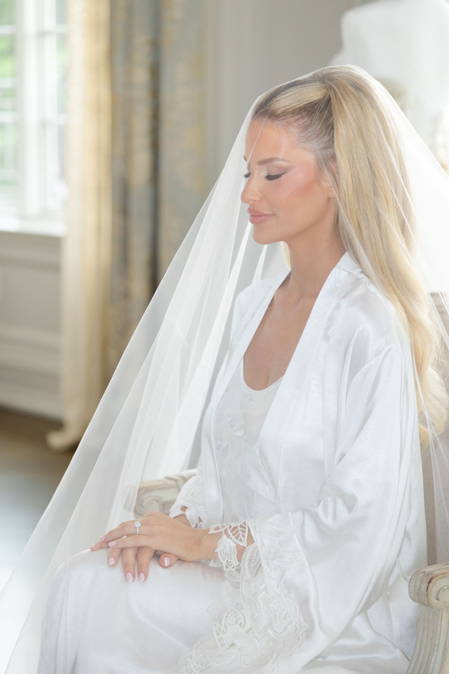 A serene bride in a white silk robe and veil sits peacefully during wedding preparations in a sunlit room.