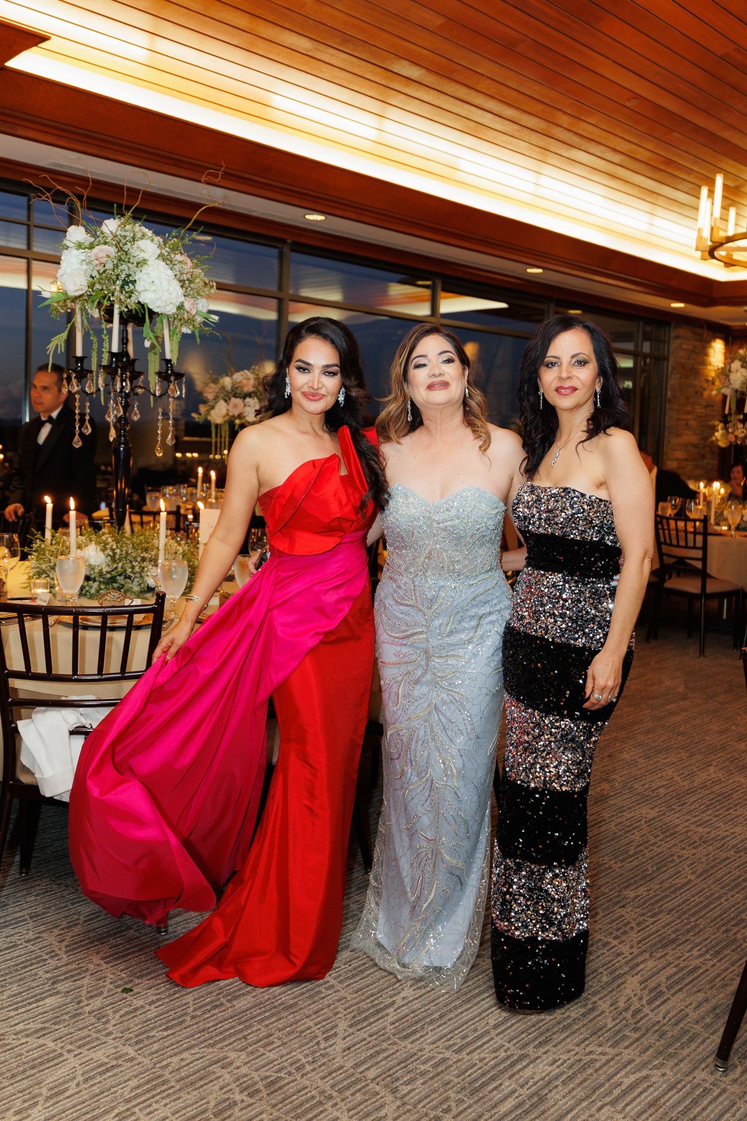 Three women in formal evening gowns - red, silver, and black striped - pose together at an elegant event venue.