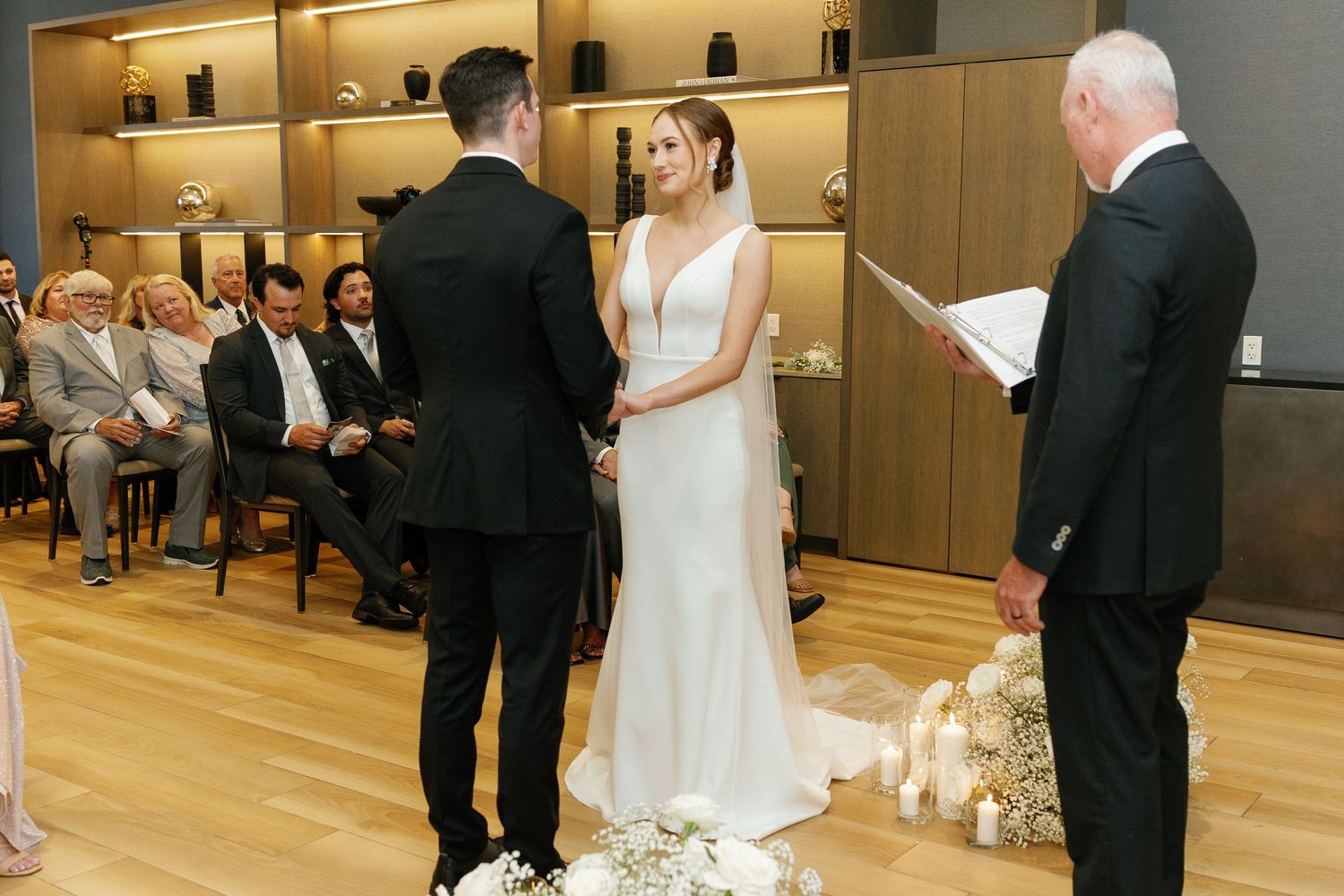 A wedding ceremony takes place in a modern venue with wooden floors and illuminated display shelves in the background.