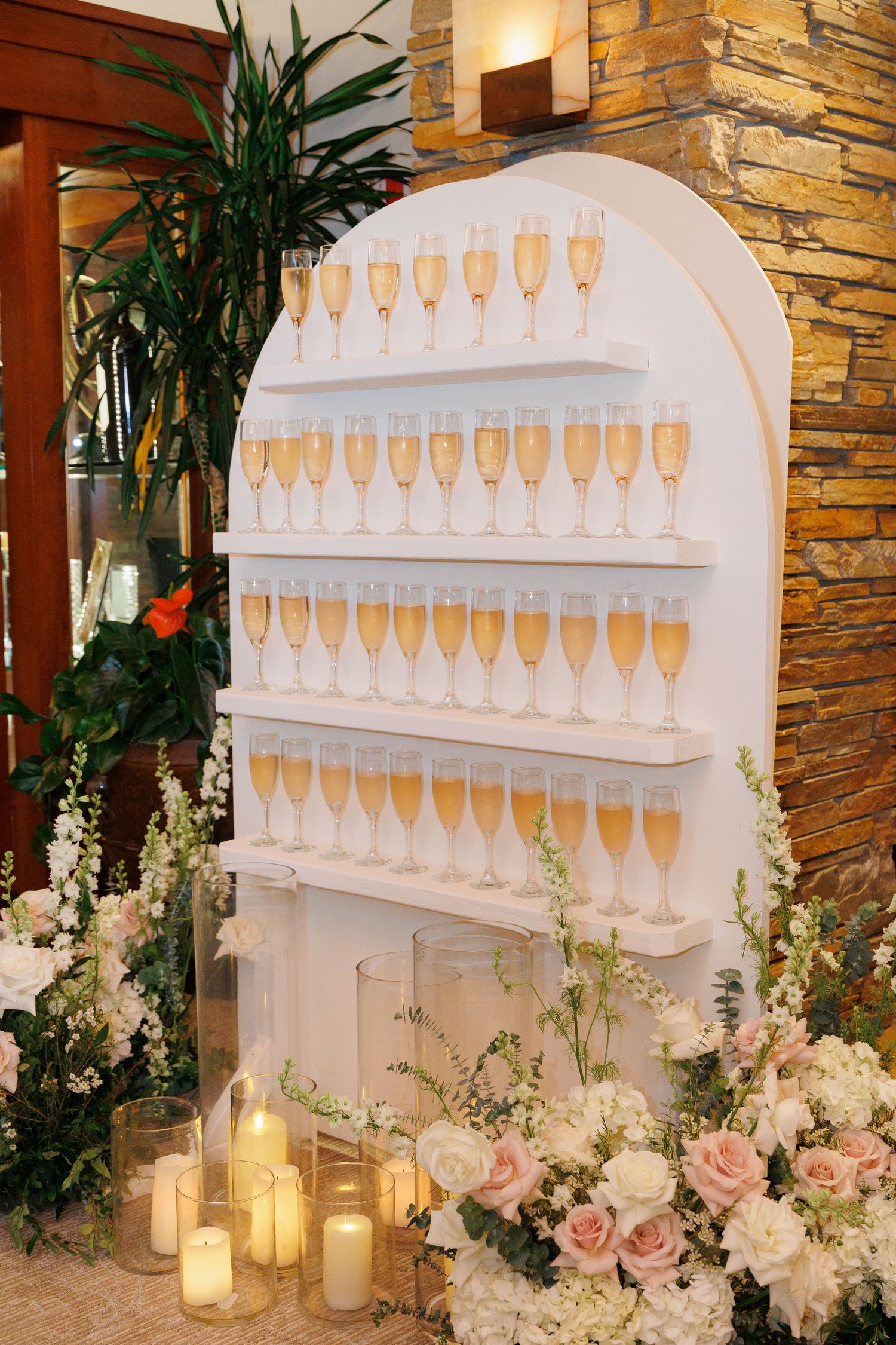 Elegant champagne wall display with white shelving, candles, and soft pink floral arrangements at an upscale event.