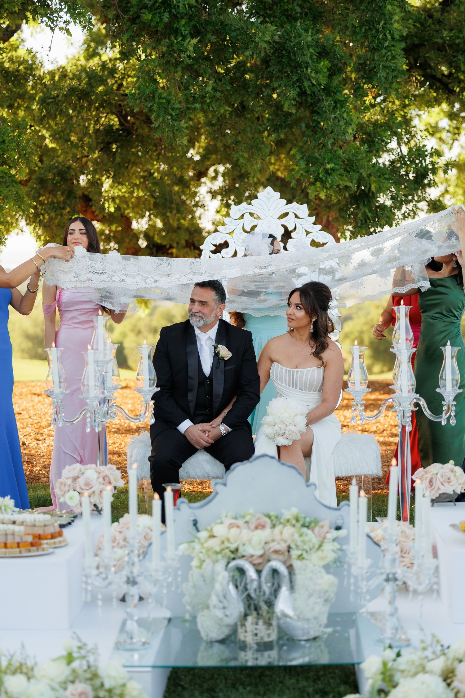 An elegant outdoor wedding reception setup features ornate white furniture and floral arrangements under lush green trees.