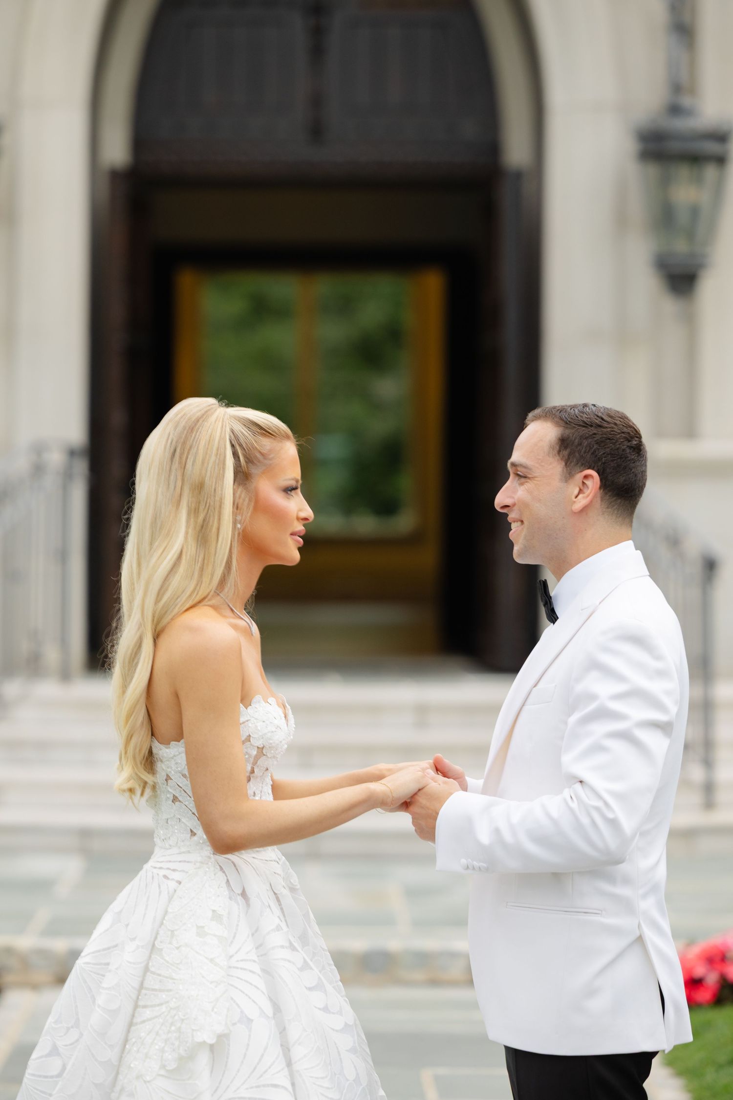 A couple in wedding attire hold hands and gaze at each other in front of an elegant building entrance.