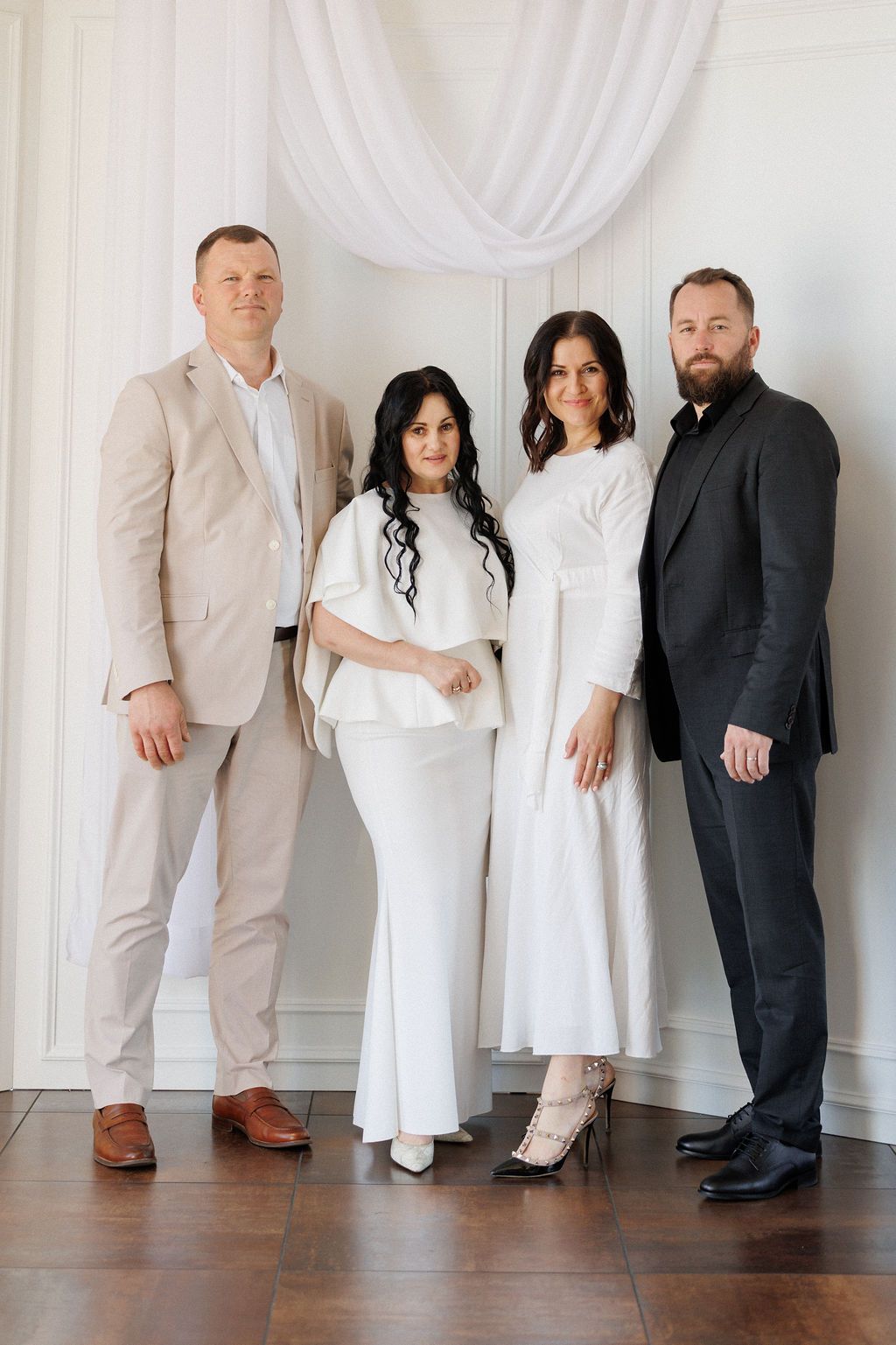 A formal group portrait of four people wearing elegant white and dark outfits against a white draped backdrop.