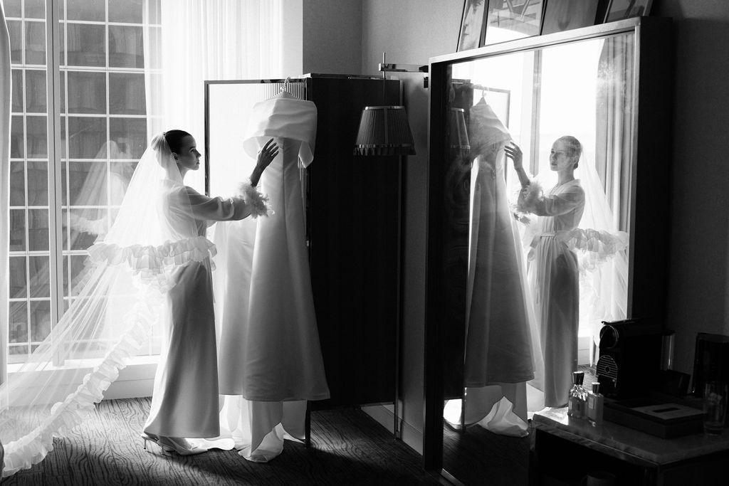 Black and white photo showing wedding dress preparations with sunlit windows casting dramatic shadows.