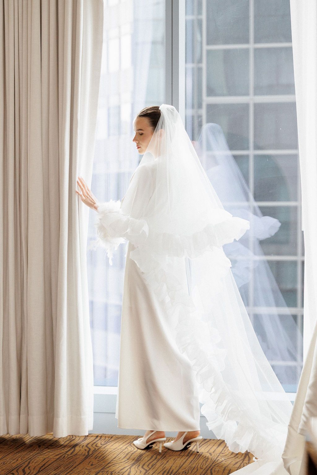 A person in flowing white fabric dances gracefully by a window with sheer curtains in a bright room.