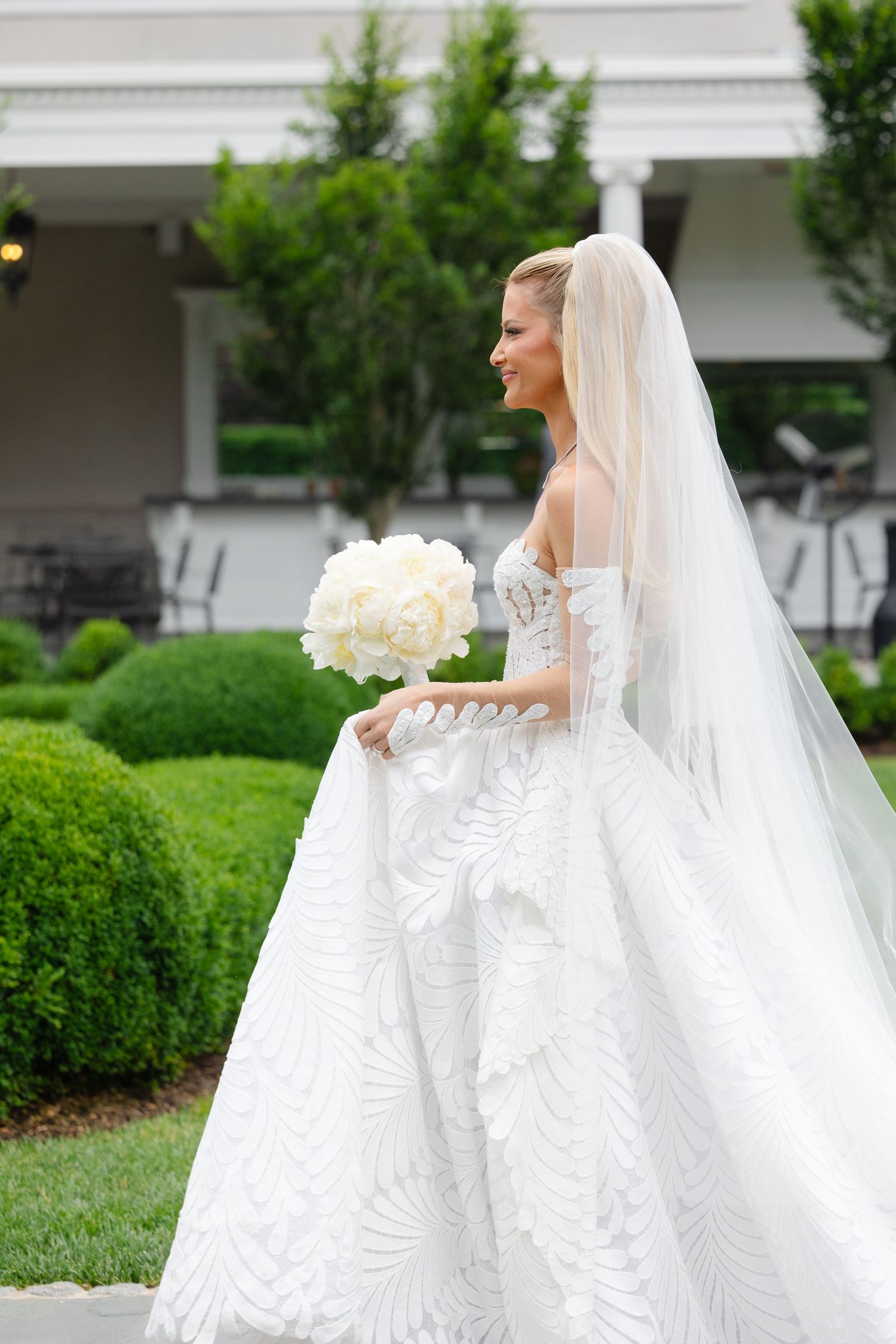 A bride in a flowing white wedding gown and veil stands in a garden setting with manicured hedges and greenery.