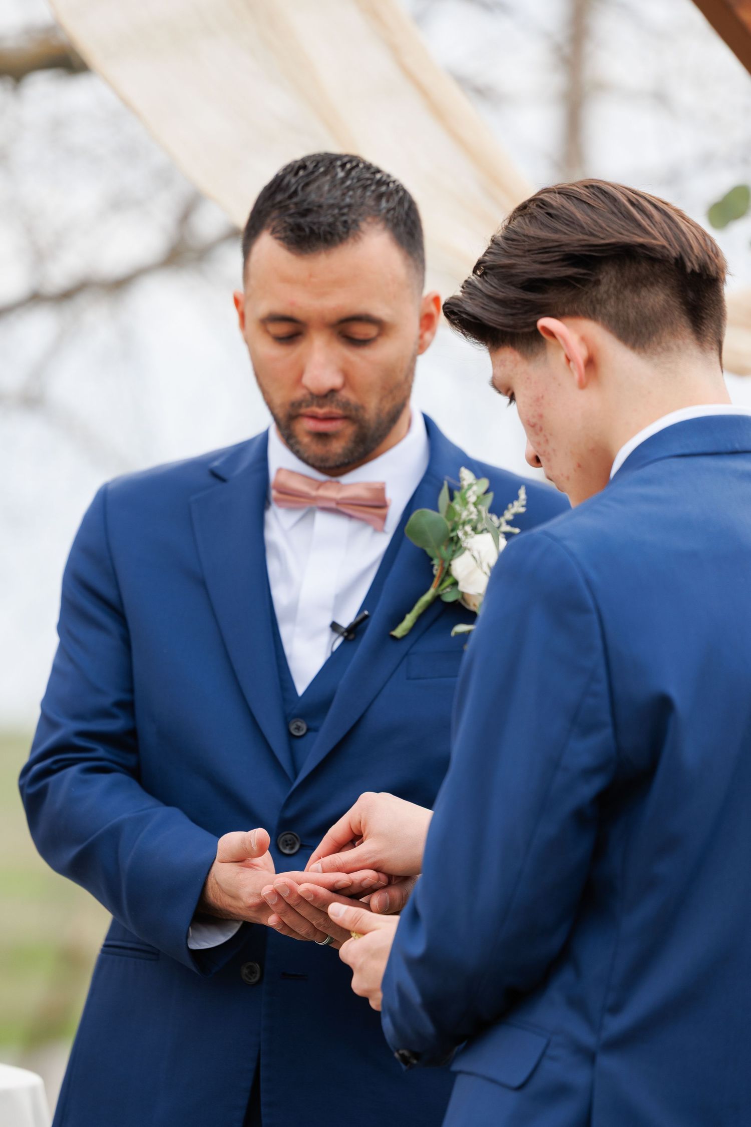 Two people in navy blue suits exchange rings during an outdoor wedding ceremony.