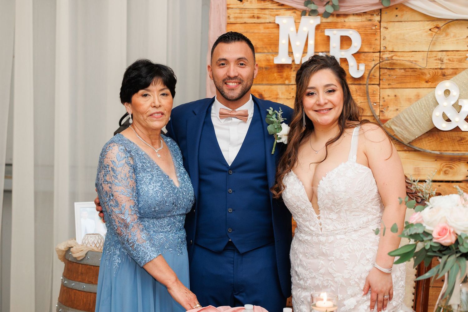 Wedding party members pose together in front of rustic wooden wall with 'MR &' signage and floral decor.