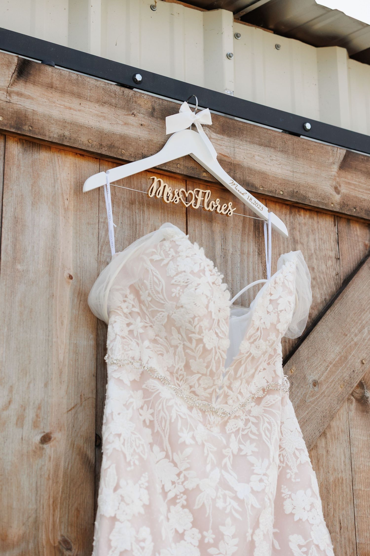 White lace wedding dress with intricate floral details hangs on wooden hangers against rustic barn wood wall.