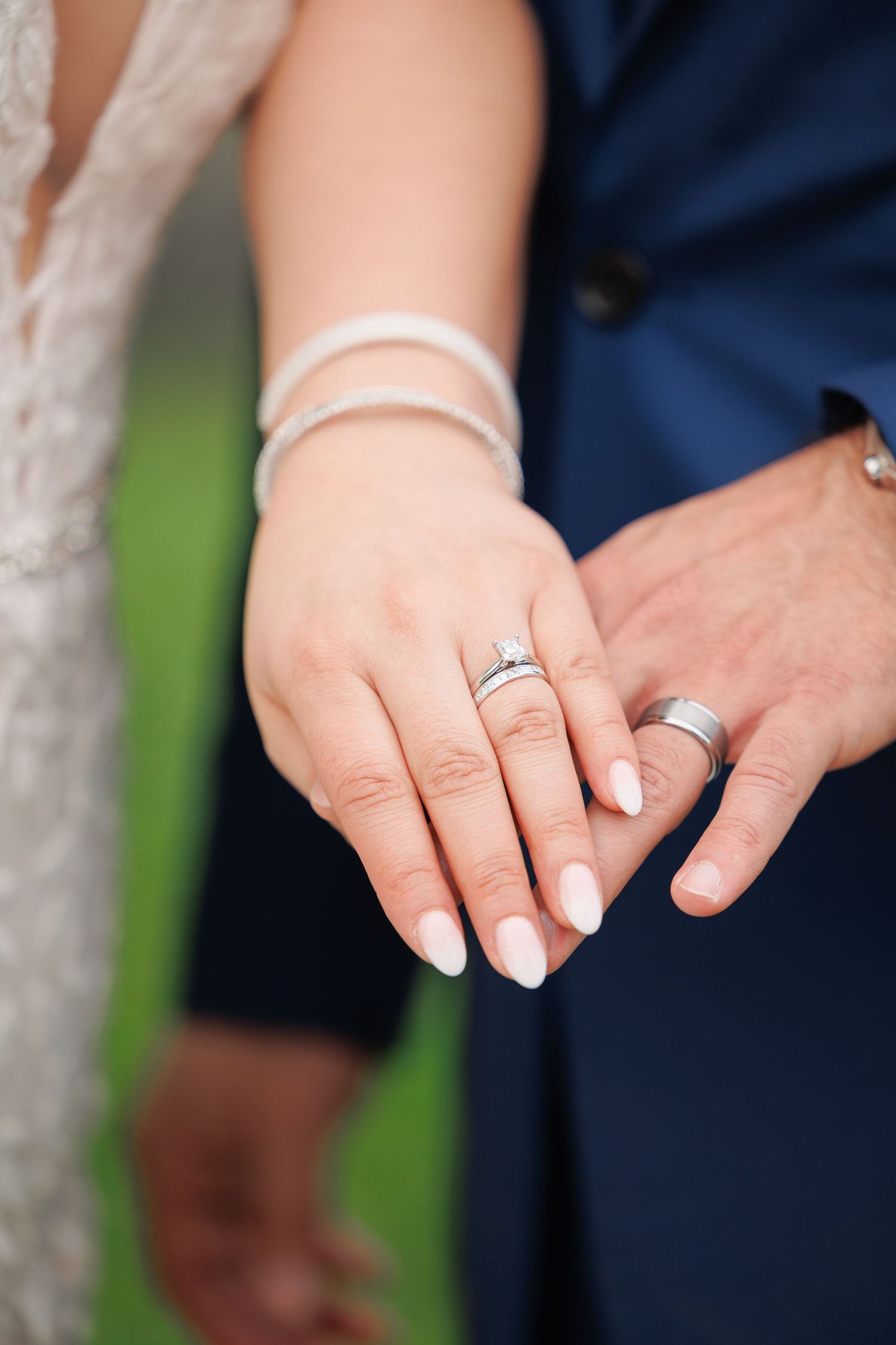 Close-up photo of hands showing wedding rings with white manicured nails against a blurred outdoor background.