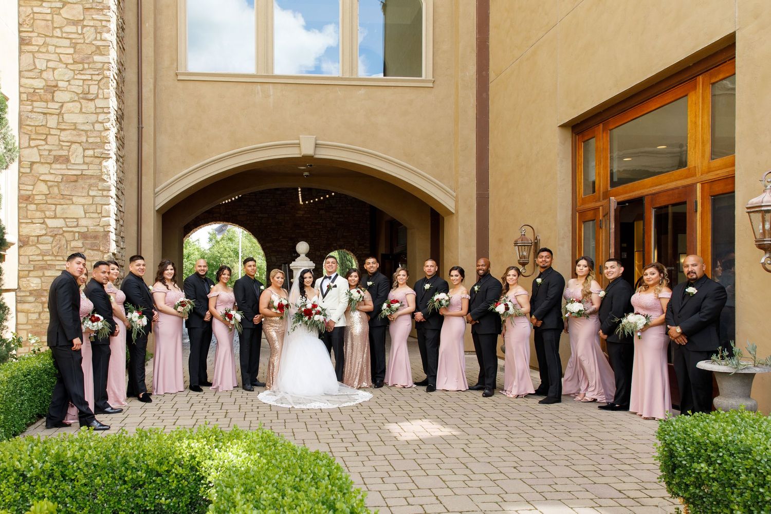 Wedding party in pink bridesmaids dresses and formal attire stands in line against stone building exterior.