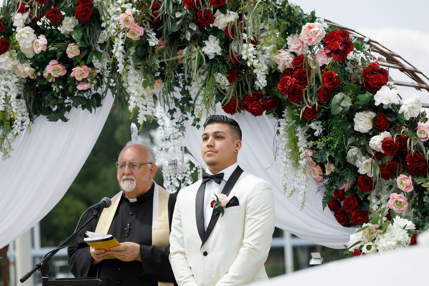 A wedding ceremony scene with red and white floral arrangements and sheer white drapes decorating the outdoor venue.