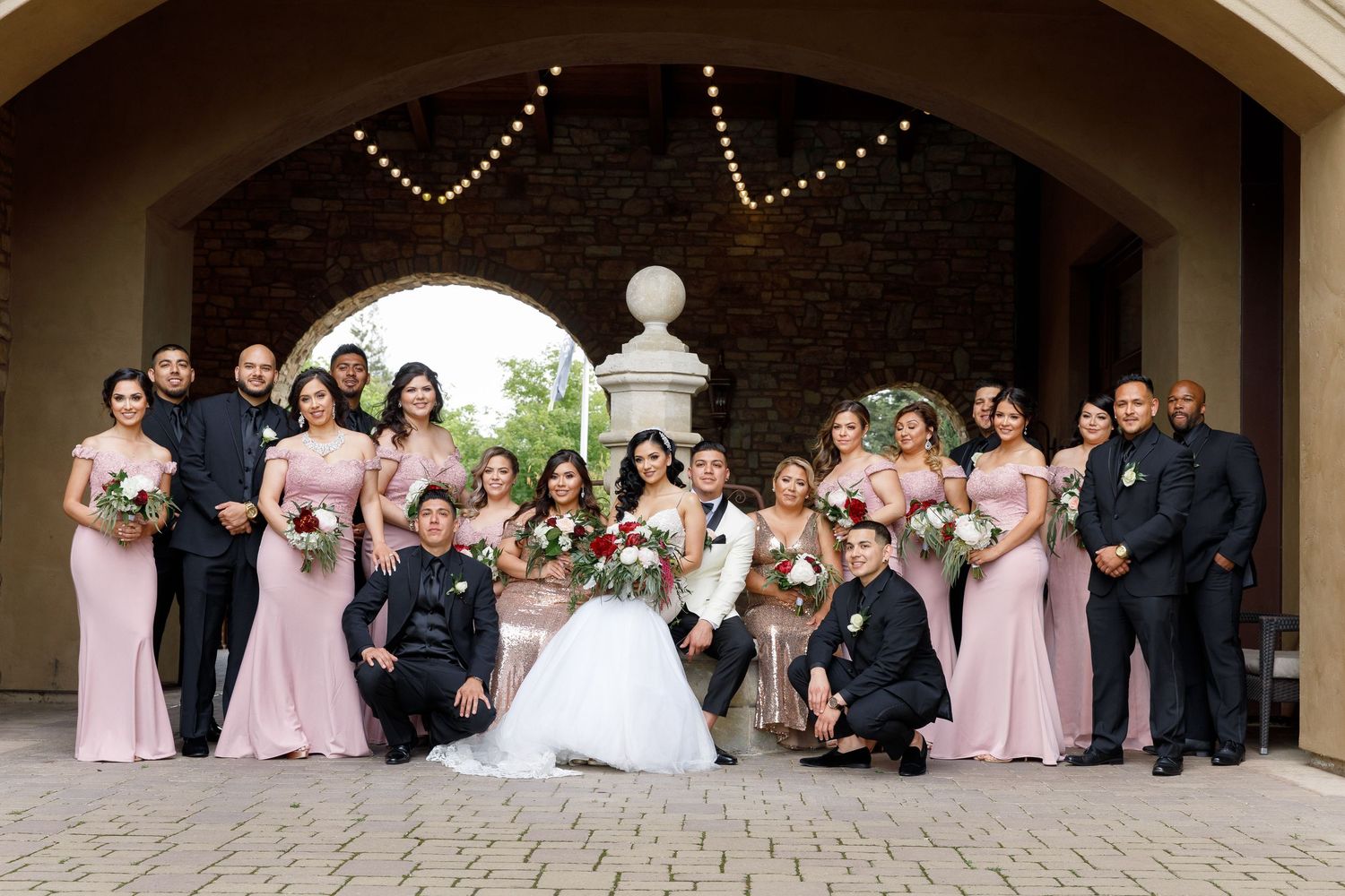 Wedding party in formal attire poses together under covered outdoor archway in elegant photo lineup.