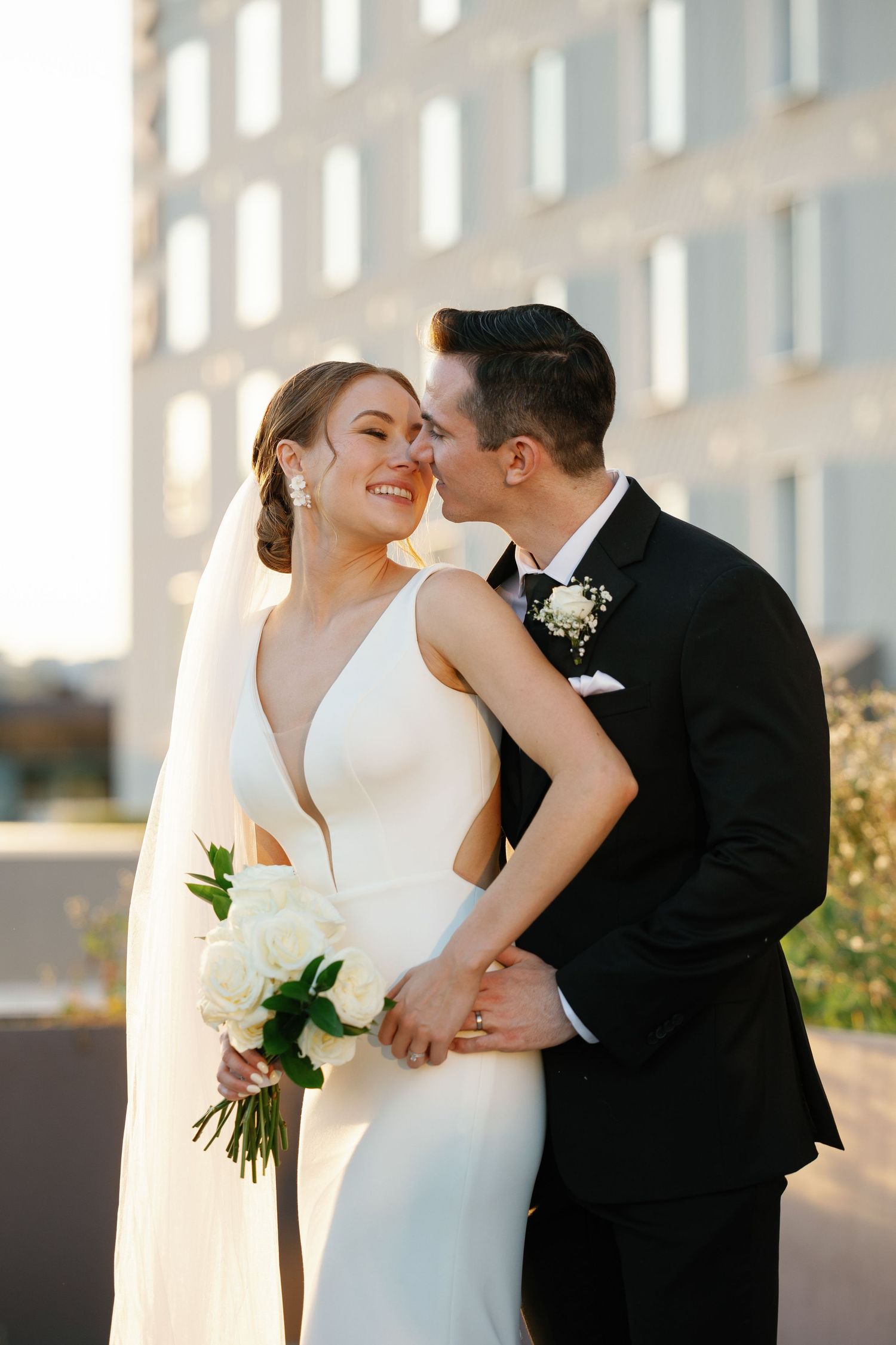 A romantic wedding couple shares tender kisses in a series of candid moments captured against a sunlit urban backdrop.