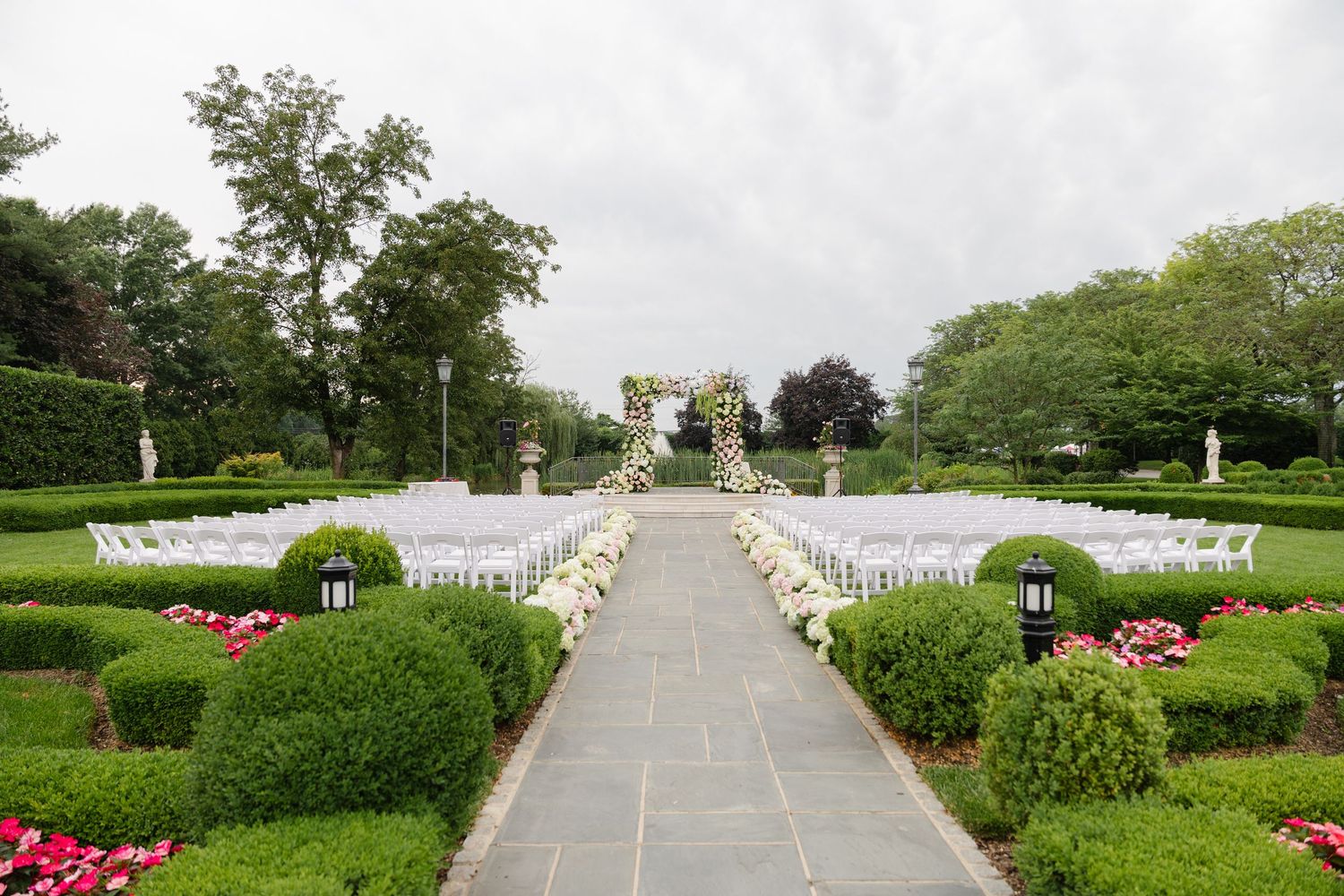A panoramic view of a formal garden with white columns, manicured hedges, and pink flowers along a symmetrical walkway.