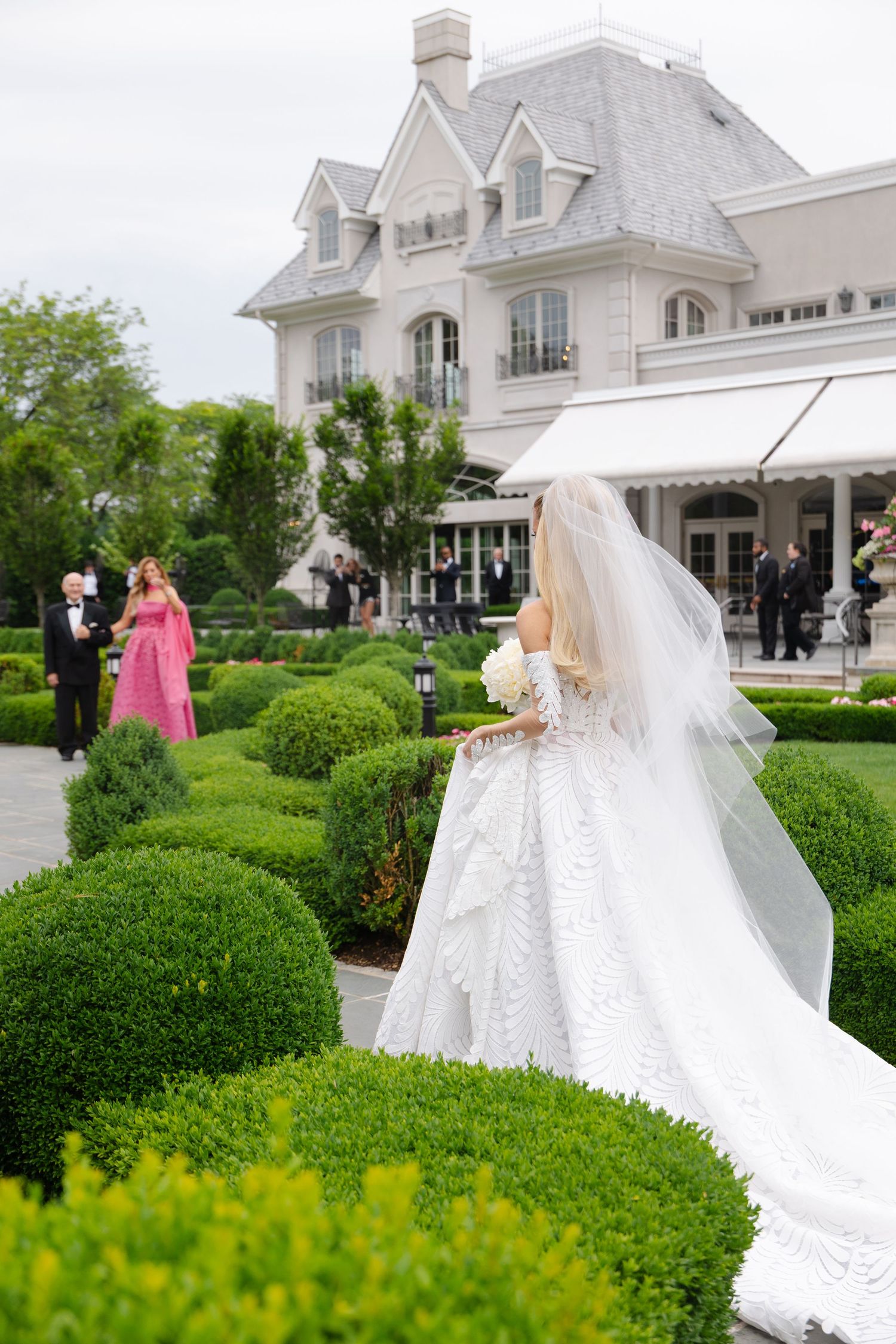 An elegant wedding celebration takes place in a manicured garden outside a grand white mansion with dormers.