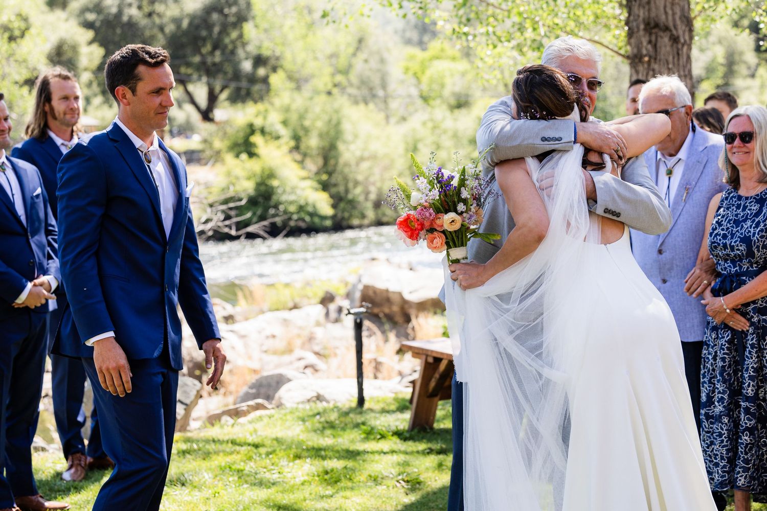 A couple shares a romantic dance outdoors at a summer wedding celebration surrounded by guests and natural scenery.