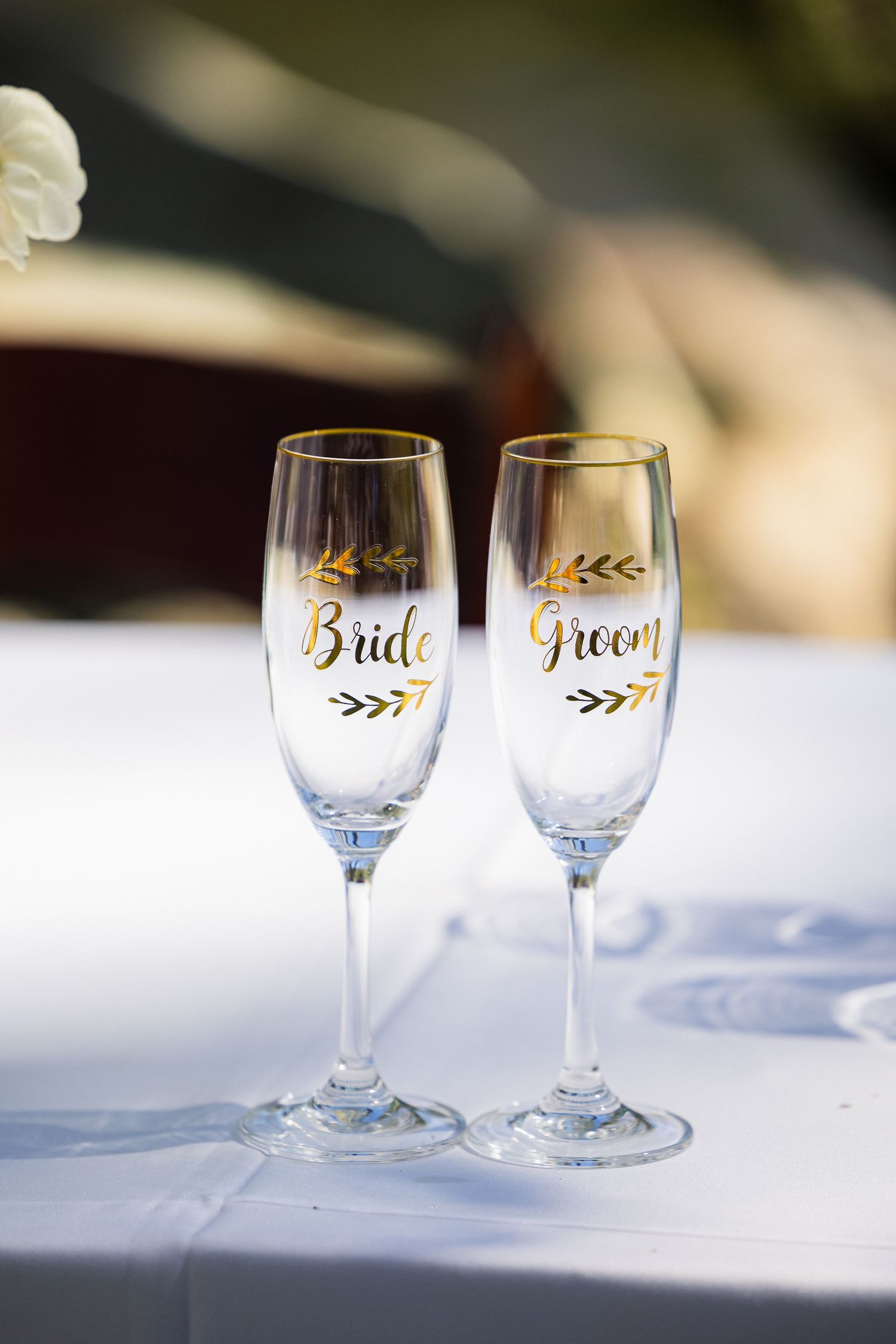 Two elegant wine glasses with gold 'Bride' and 'Groom' lettering and delicate leaf designs sit on a white tablecloth.