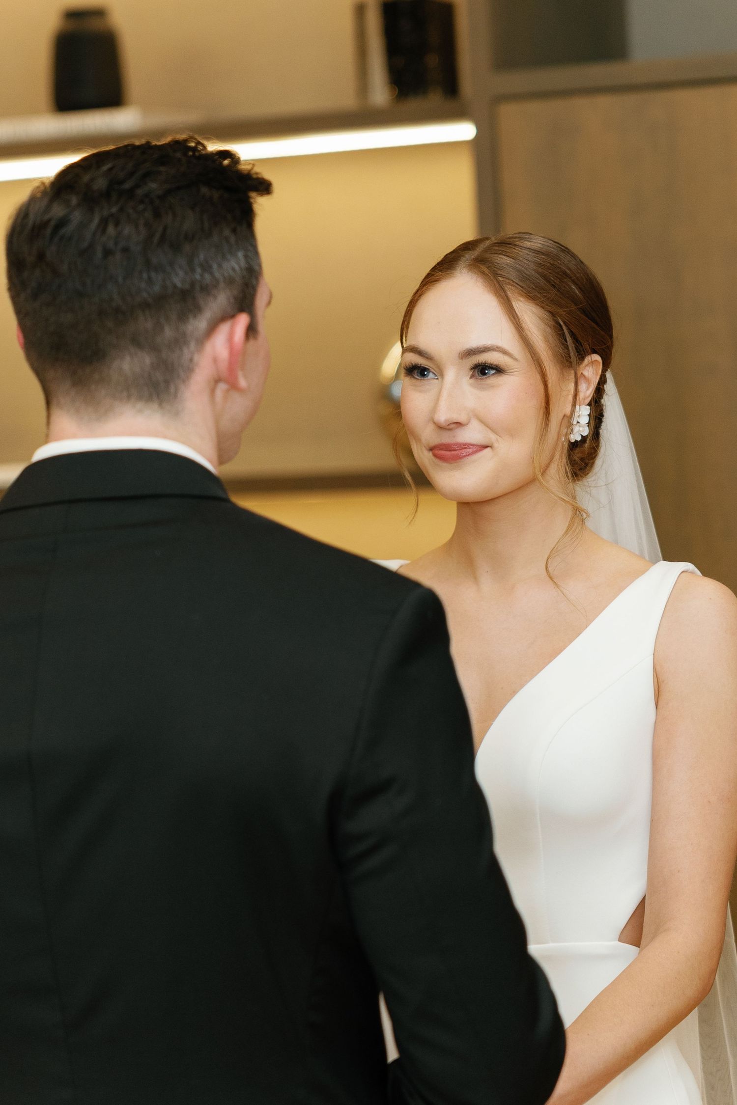 Bride in elegant white sleeveless wedding dress smiles while groom in black suit stands with back turned.