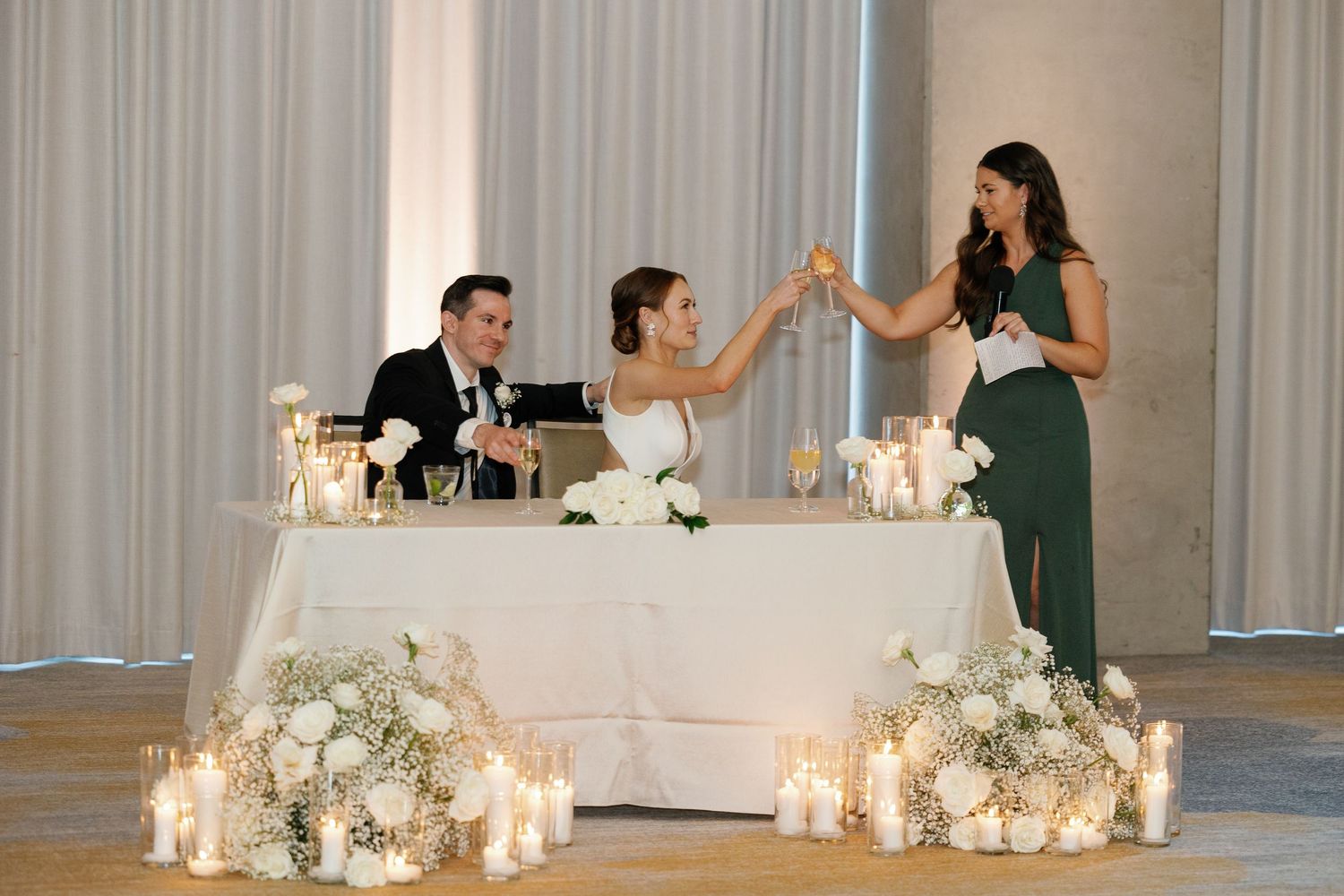 Wedding party members raise their glasses in a toast at an elegant reception with candlelit white floral decorations.