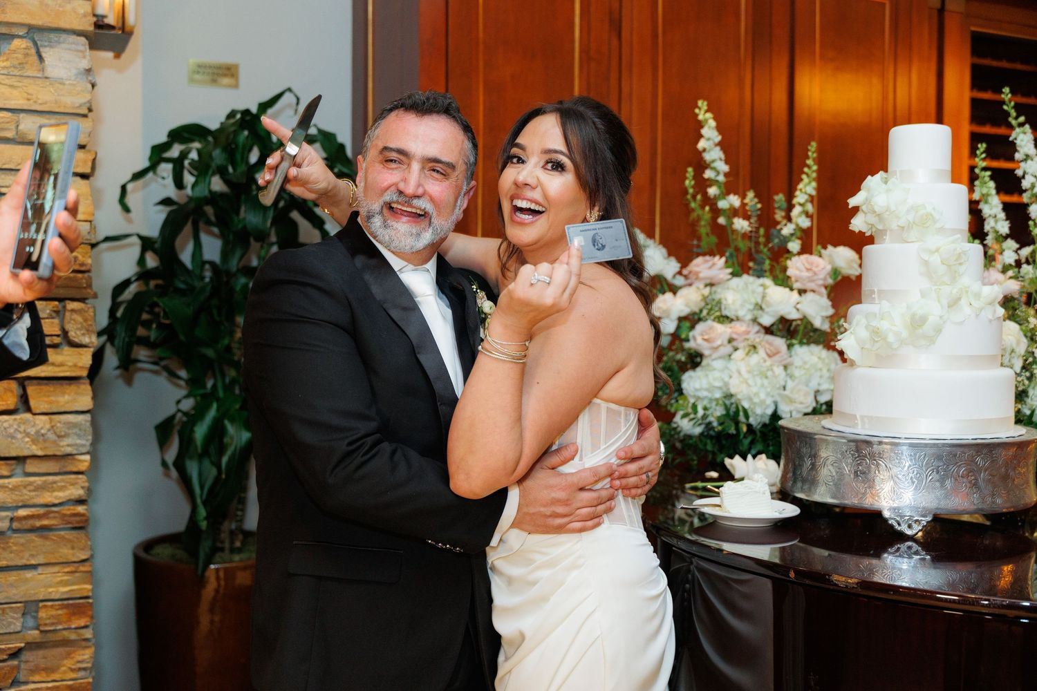 A joyful wedding celebration with people laughing and dancing near an elegant white tiered cake at a reception venue.