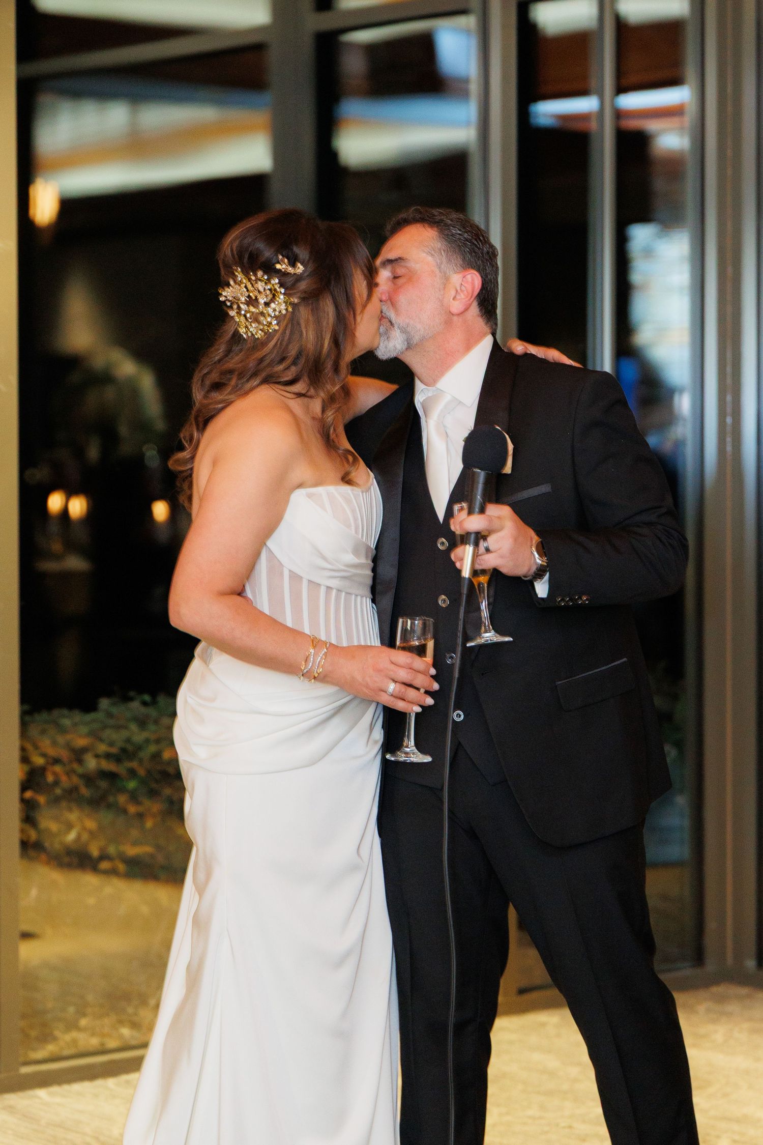 A bride in a white gown and groom in black tuxedo share a romantic moment while holding champagne glasses at their reception.