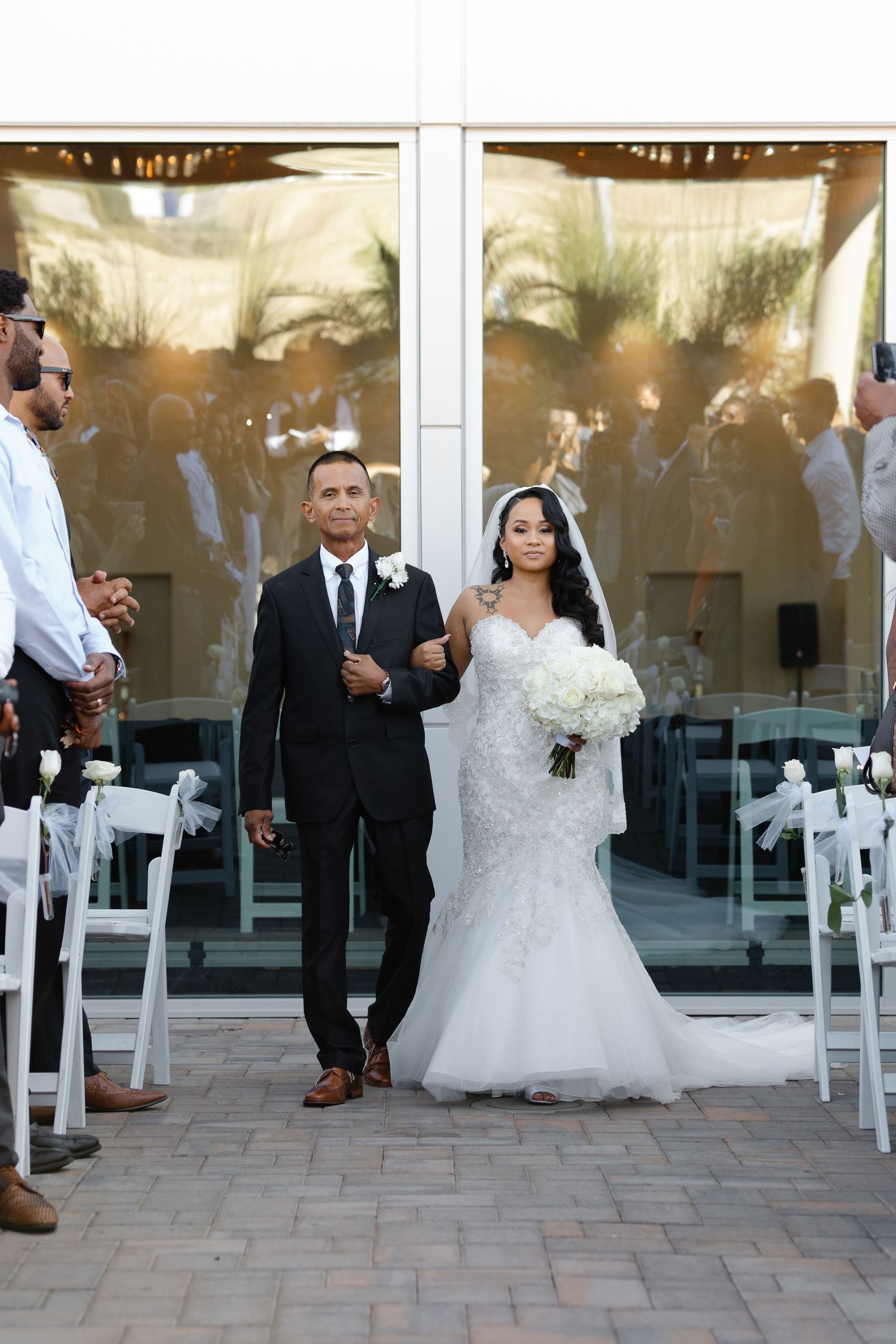 A beautiful wedding ceremony at an elegant venue with white chairs lining the aisle and glass windows in the background.