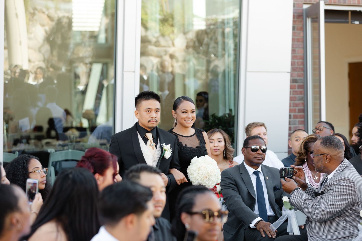 People in formal attire socialize and mingle outside a building during what looks like a special occasion.