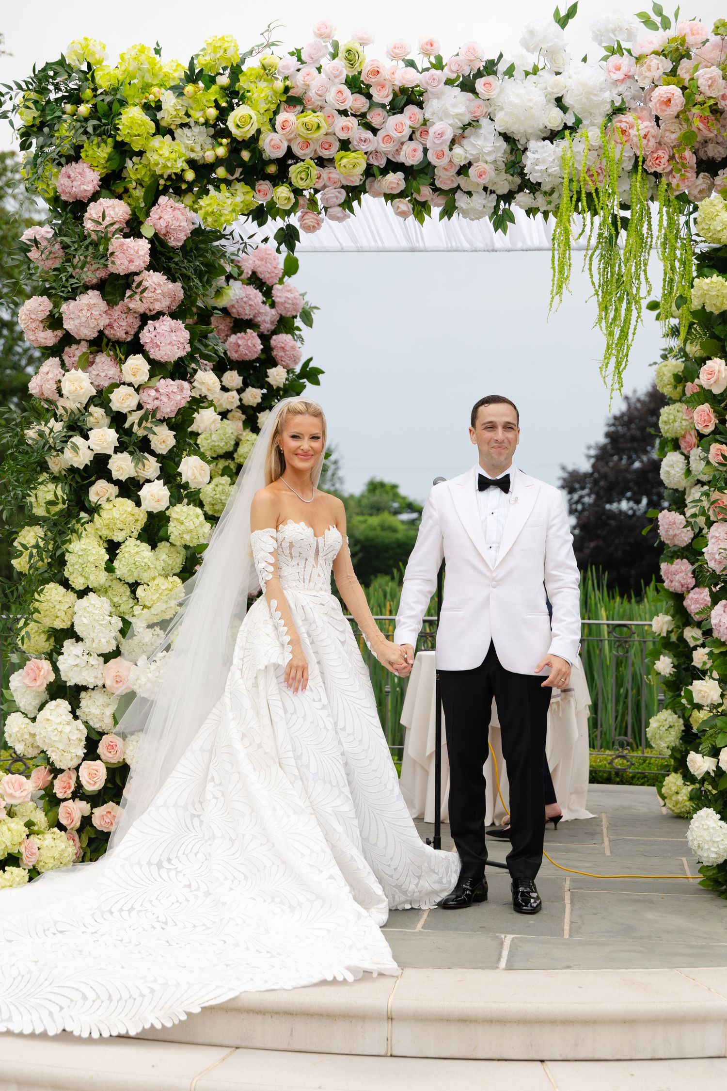 Bride and groom holding hands under a floral arch adorned with pink, white, and green flowers at an outdoor wedding.
