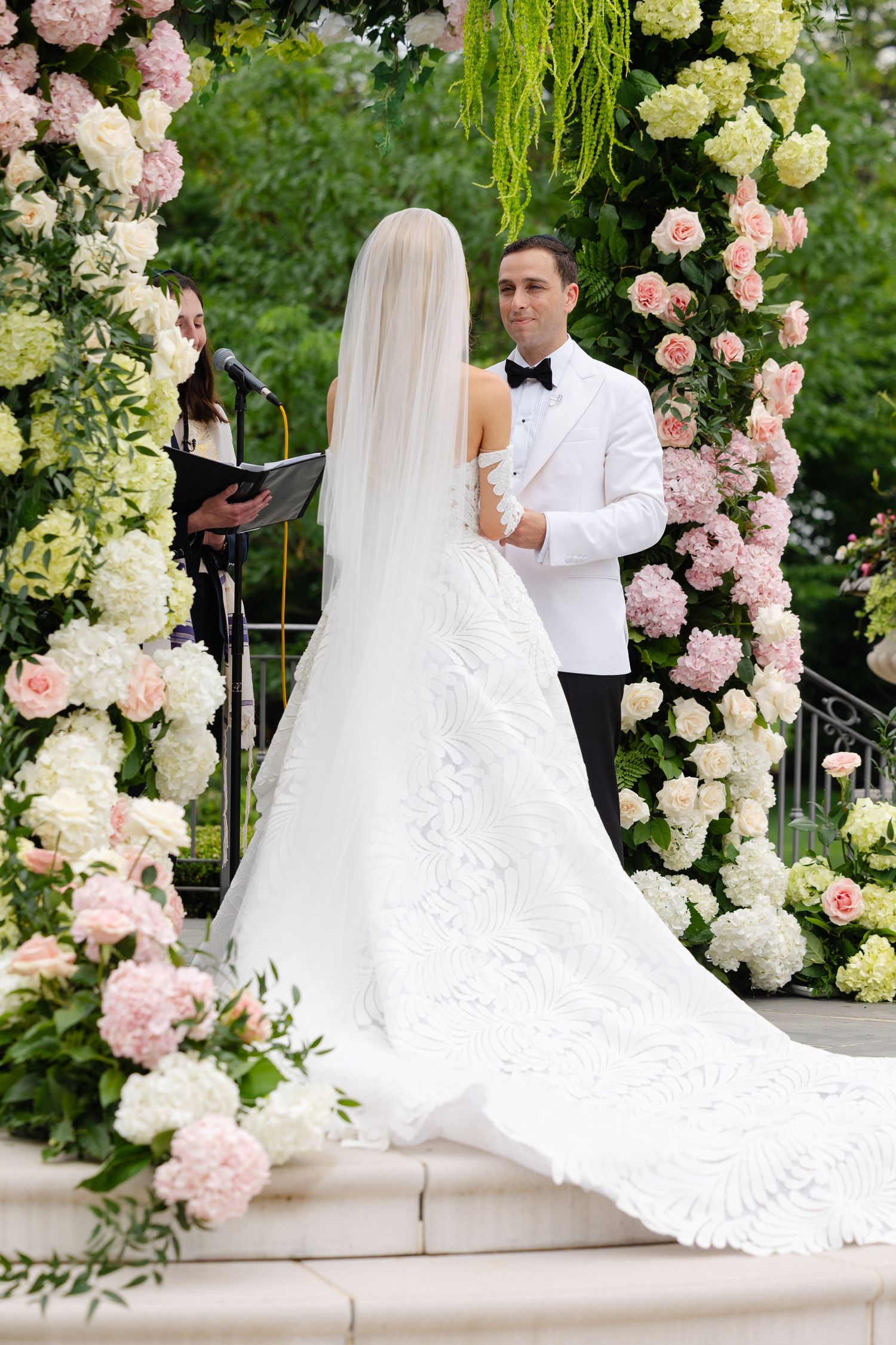 Bride and groom exchange vows at an outdoor ceremony beneath a floral arch of pink and white hydrangeas and roses.