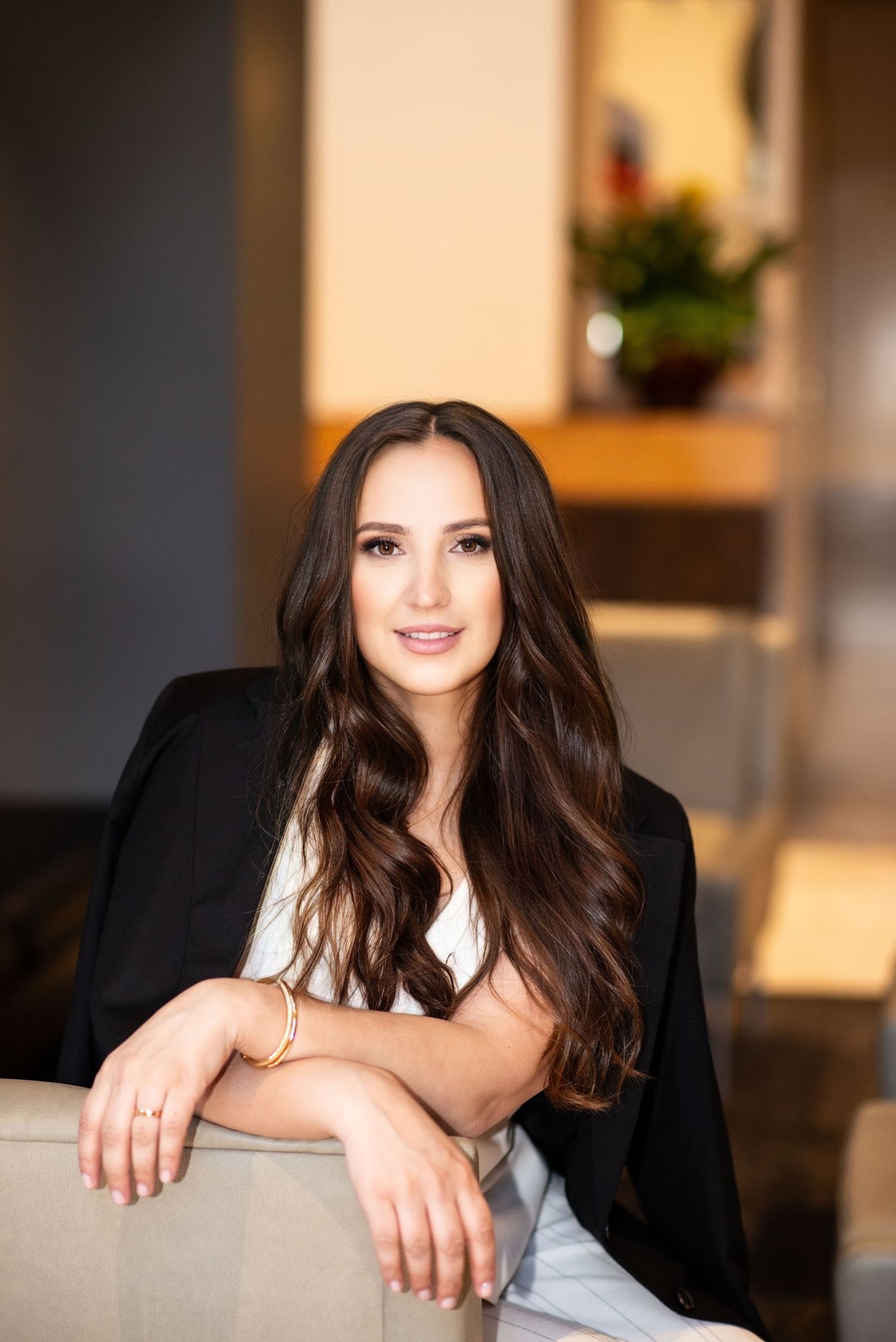 A professional portrait of a woman in a black blazer sitting in an elegant interior setting.