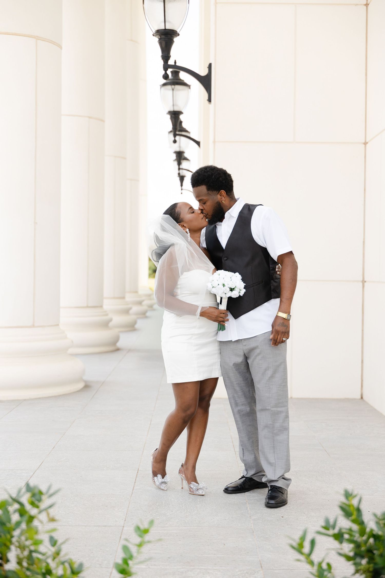 A newlywed Black couple shares a kiss, bride in white mini dress with veil holding bouquet, groom in vest and gray pants.