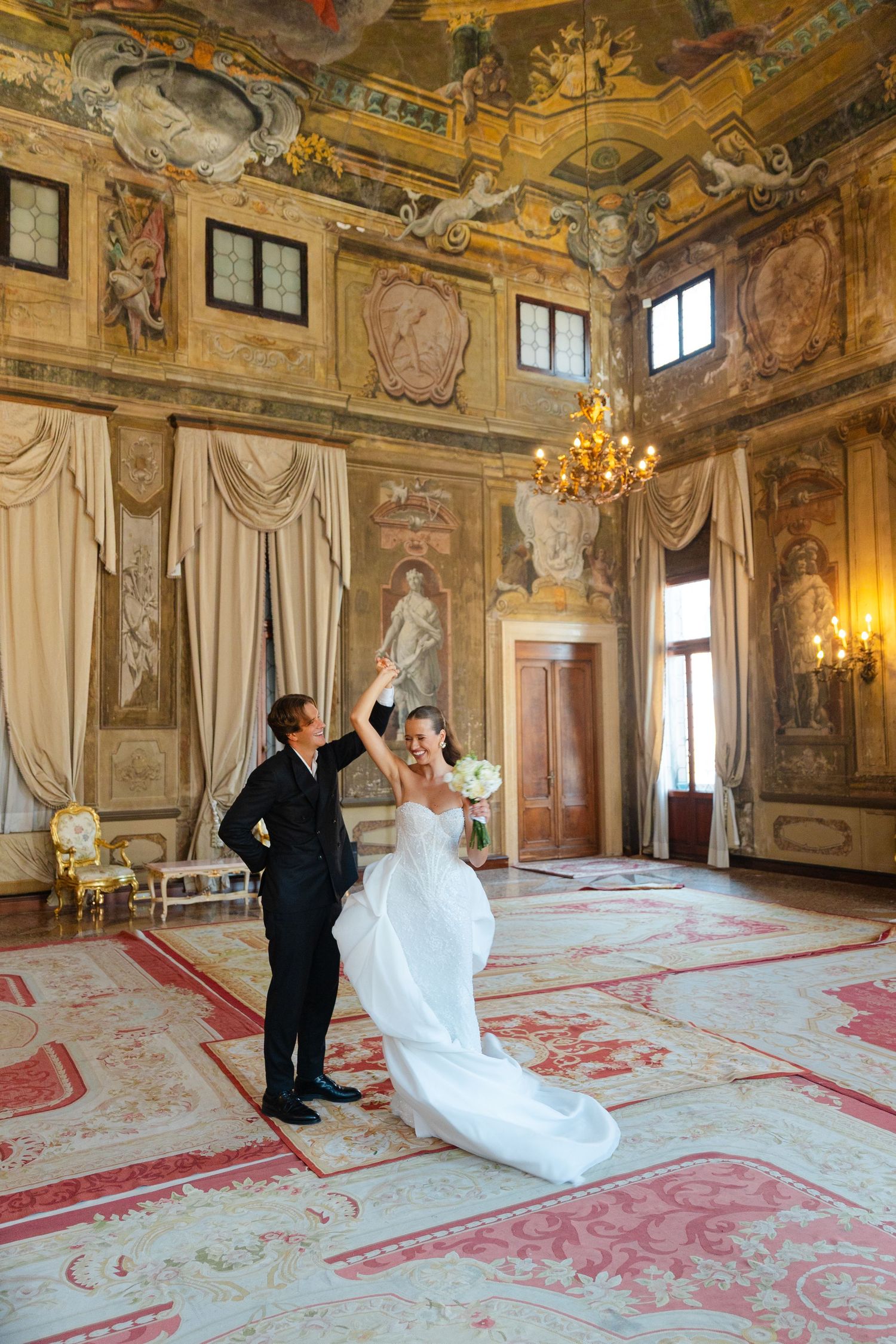 A couple dances in an ornate baroque ballroom with frescoed ceilings, crystal chandeliers and elegant red carpeting.
