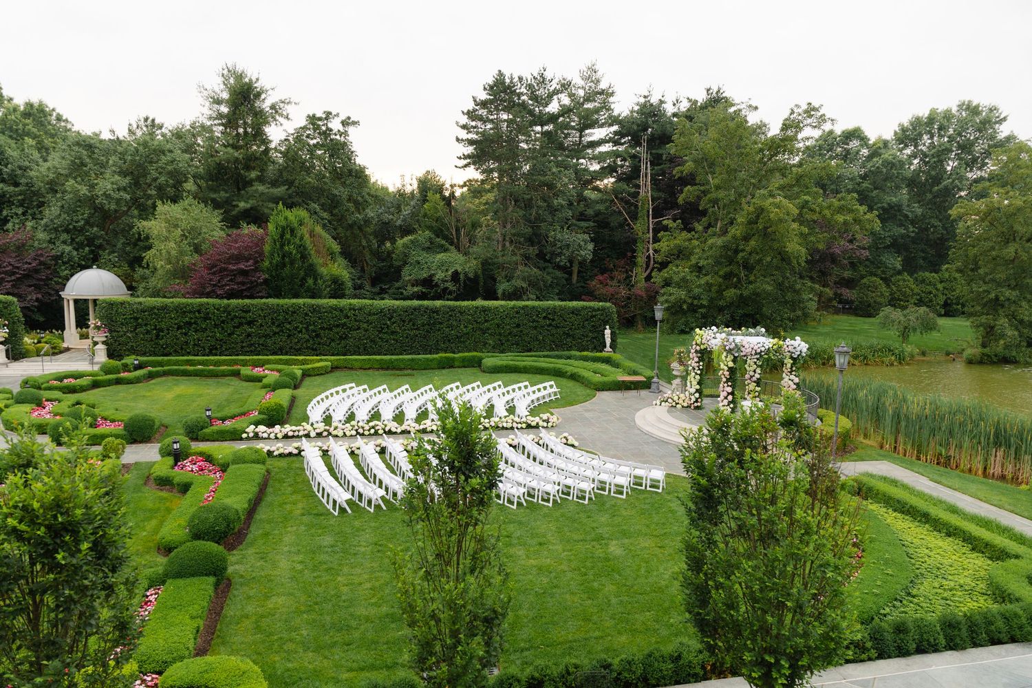 Elegant outdoor wedding ceremony setup with white chairs, floral arch, manicured hedges, and a scenic pond in a lush garden.