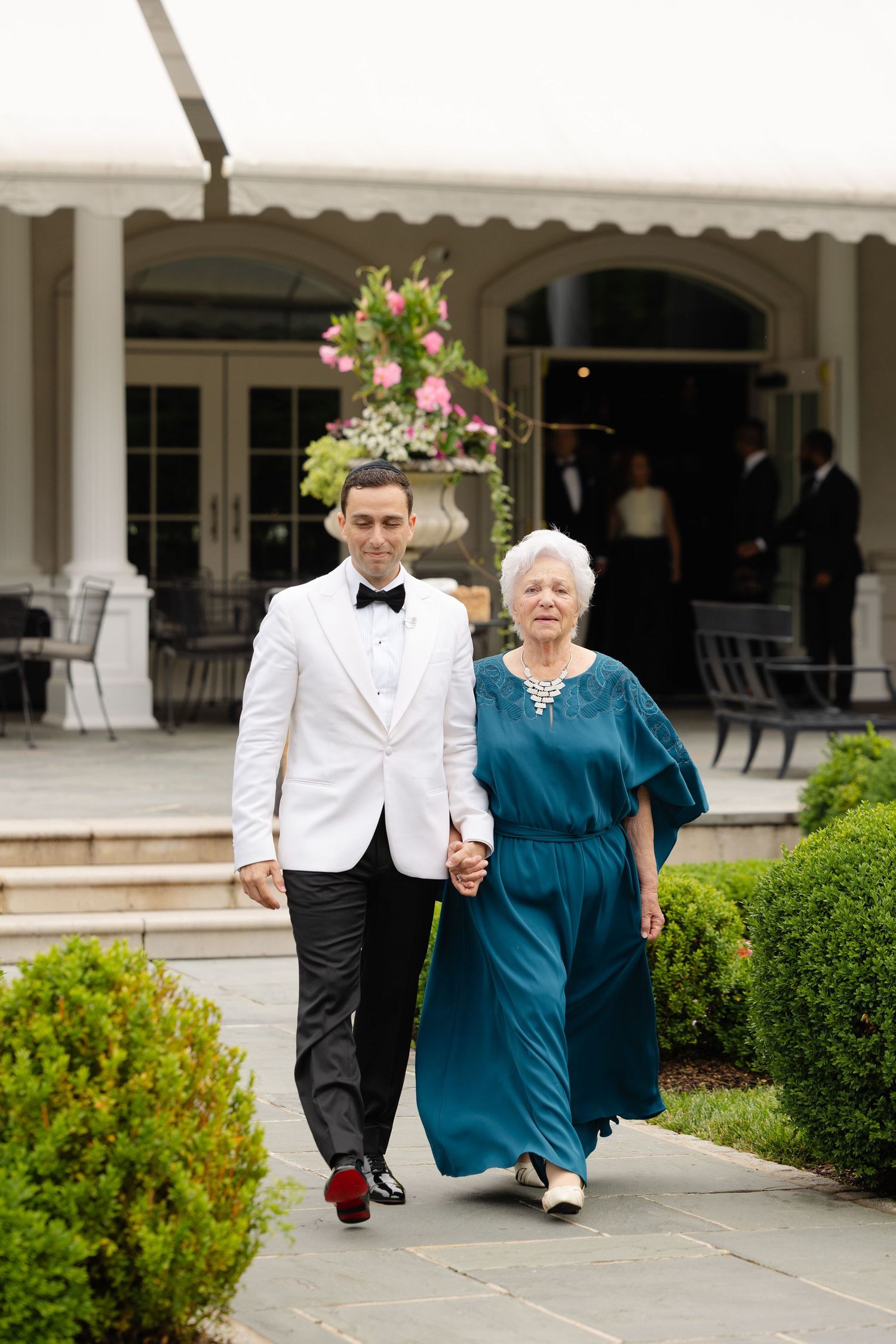 Groom in white tuxedo jacket escorts elderly woman in teal dress along garden path at elegant wedding venue.