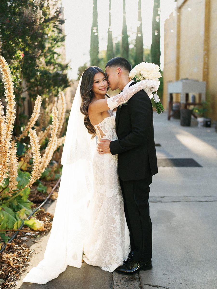 A newlywed couple embraces on their wedding day, with the bride in lace and the groom in a classic black tuxedo.