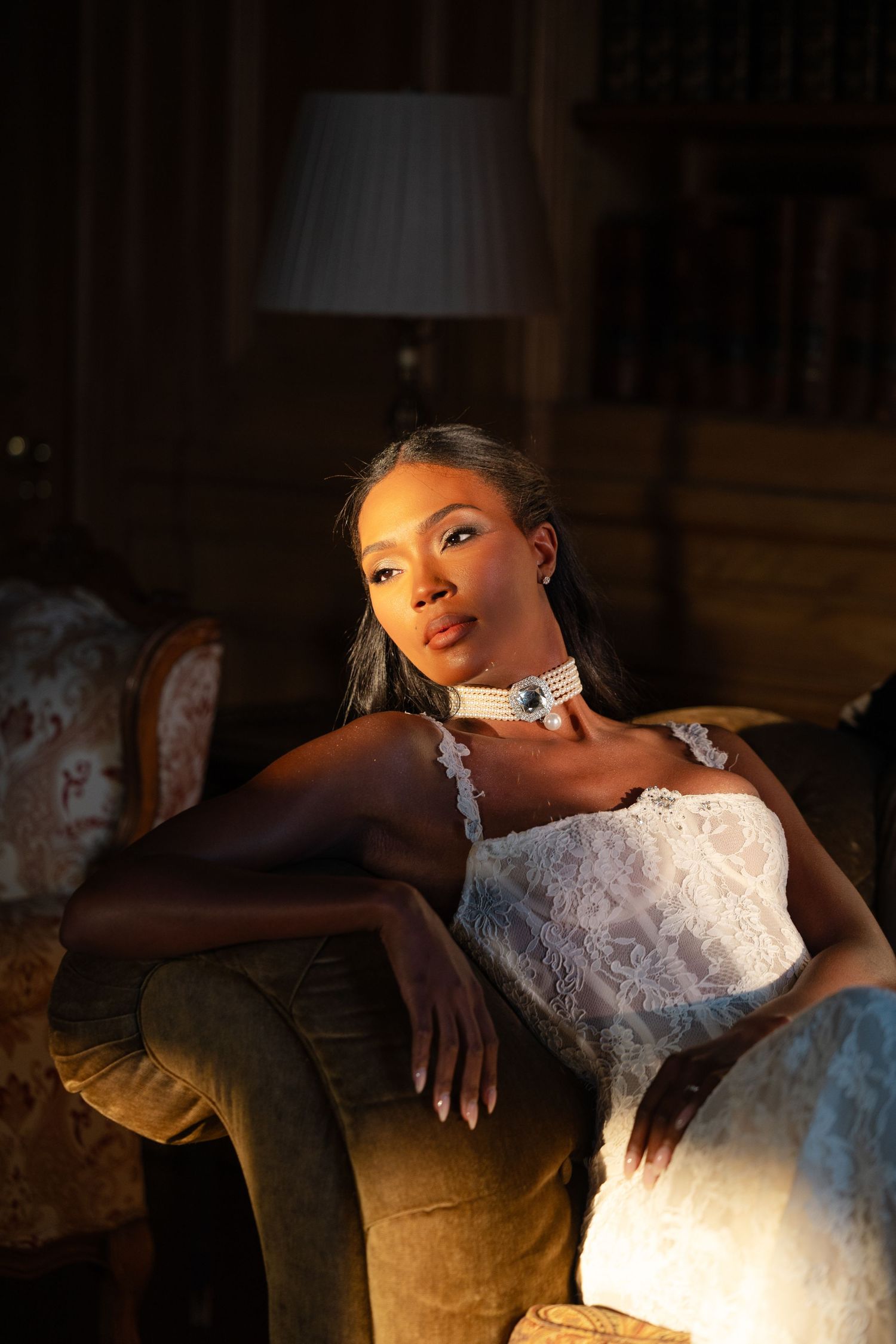 A bride in a white lace dress reclines on an elegant vintage sofa in dramatic lighting.