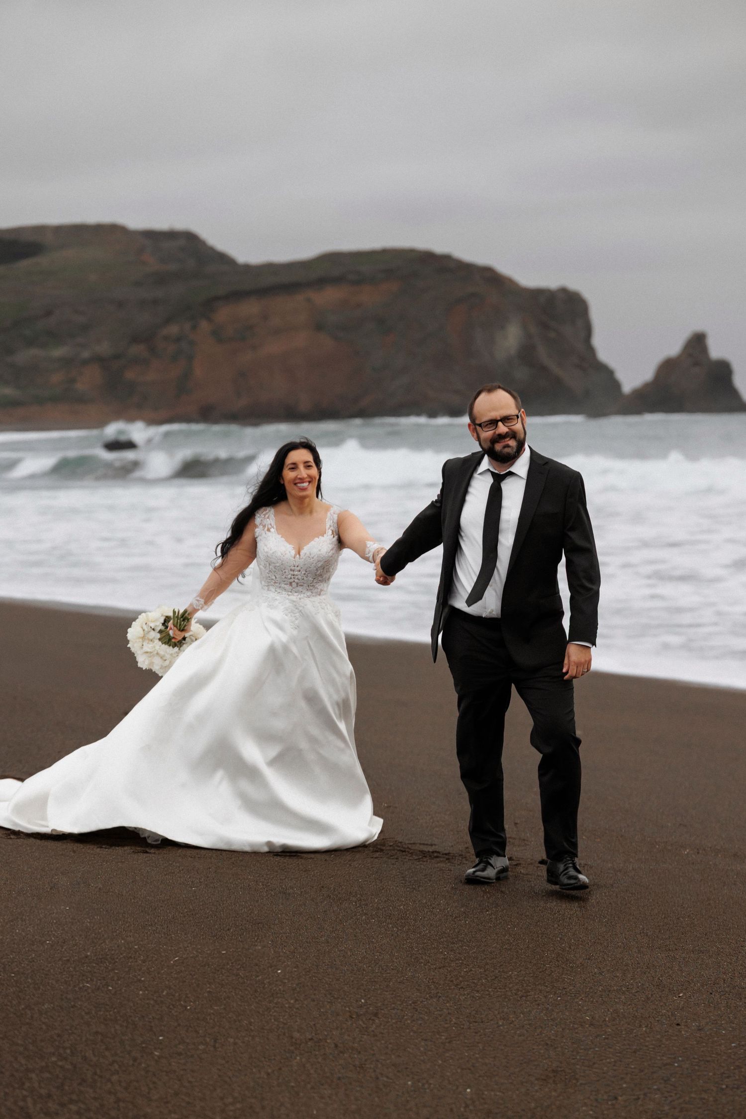 A couple in wedding attire poses playfully on a dramatic black sand beach with rocky cliffs in the background.