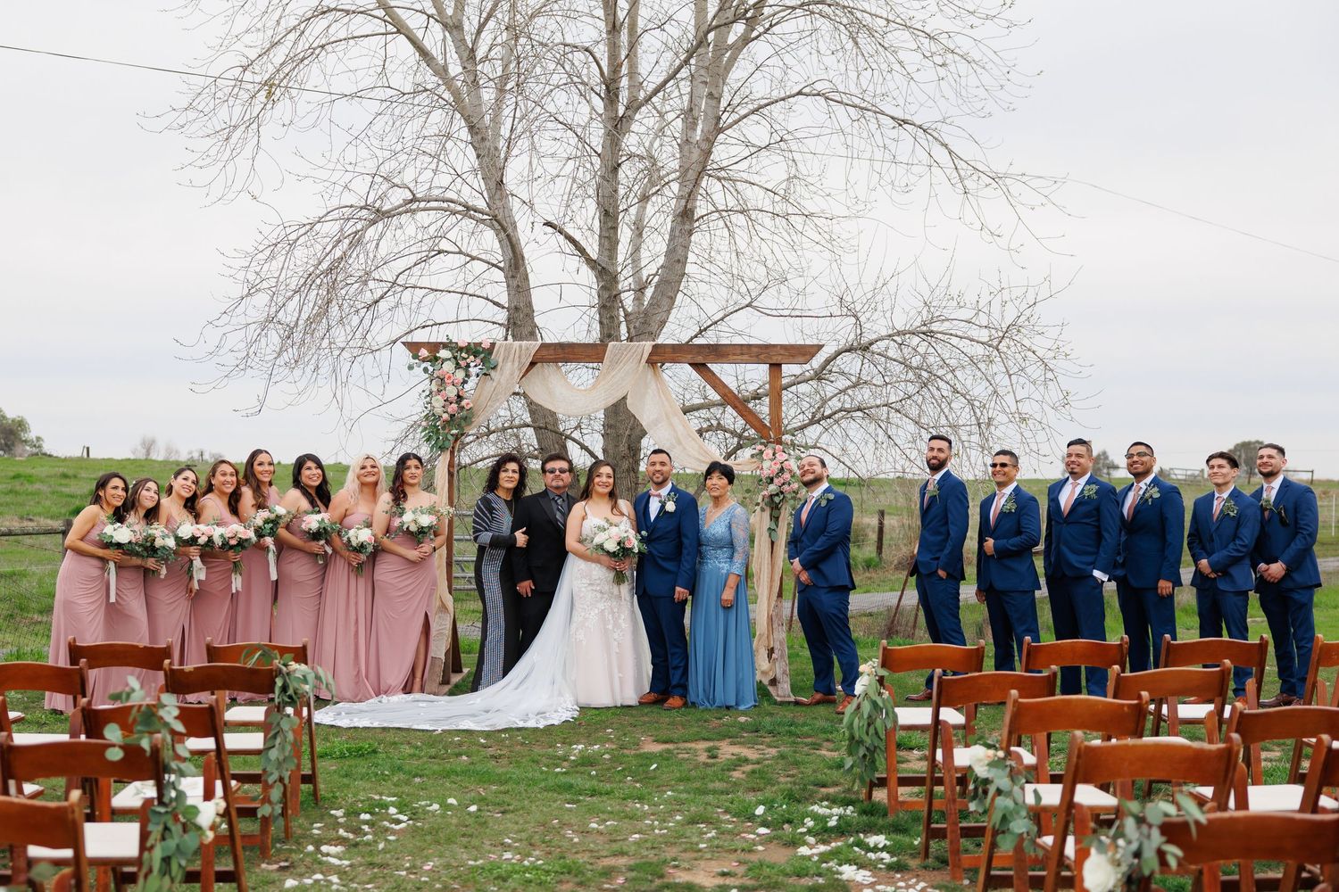 Wedding party poses outdoors in formal attire with bridesmaids in pink and blue dresses and groomsmen in navy suits.