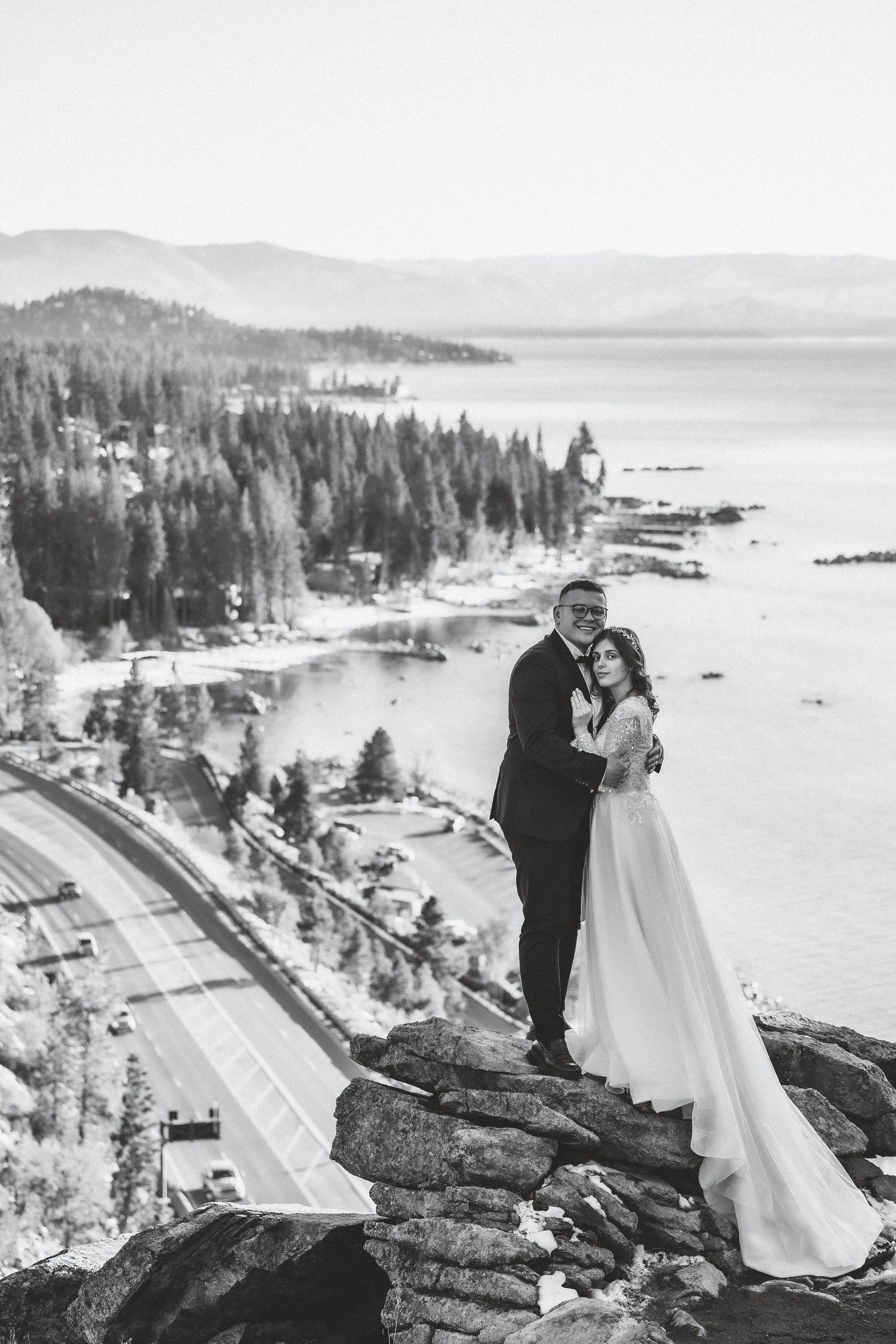 Wedding couple stands on dramatic cliff overlooking mountain lake and winding road below.